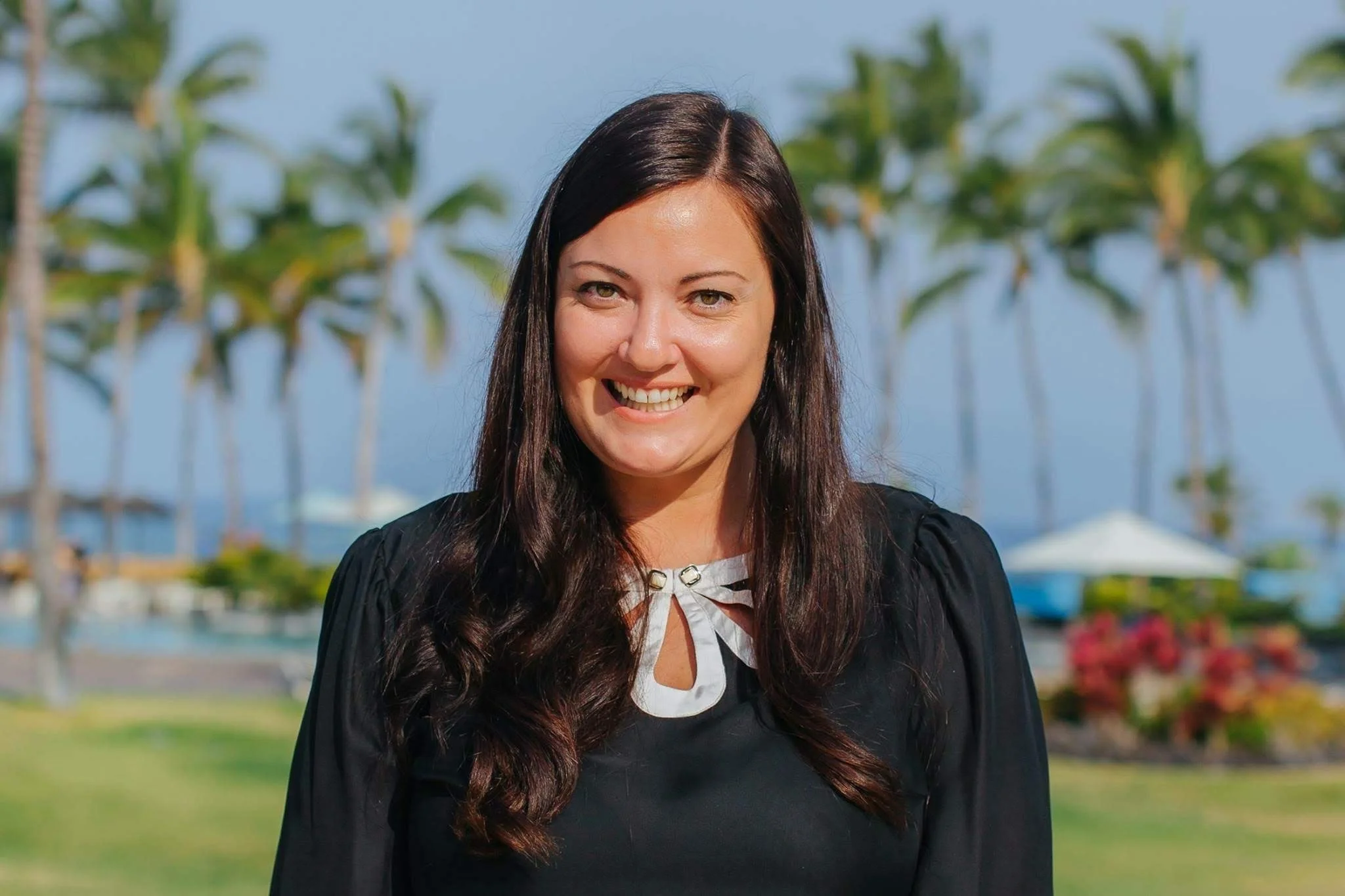 Summer Carrick on the Kohala Coast in Hawaii with long dark hair smiling outdoors near palm trees and the Pacific Ocean.
