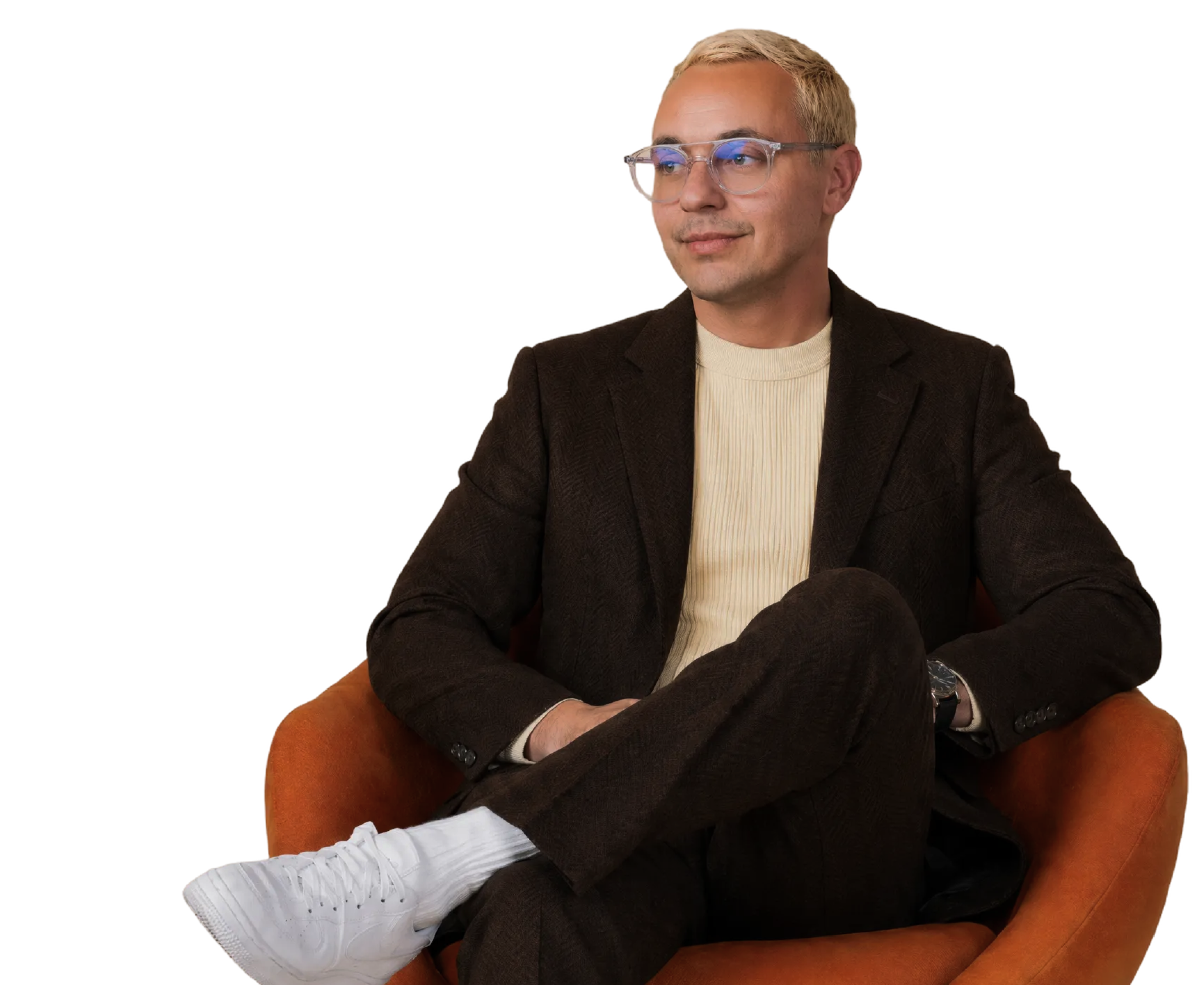 A man with short blond hair and clear glasses sits in an orange chair, wearing a black blazer and a cream turtleneck, with his arms crossed and looking slightly to the side.
