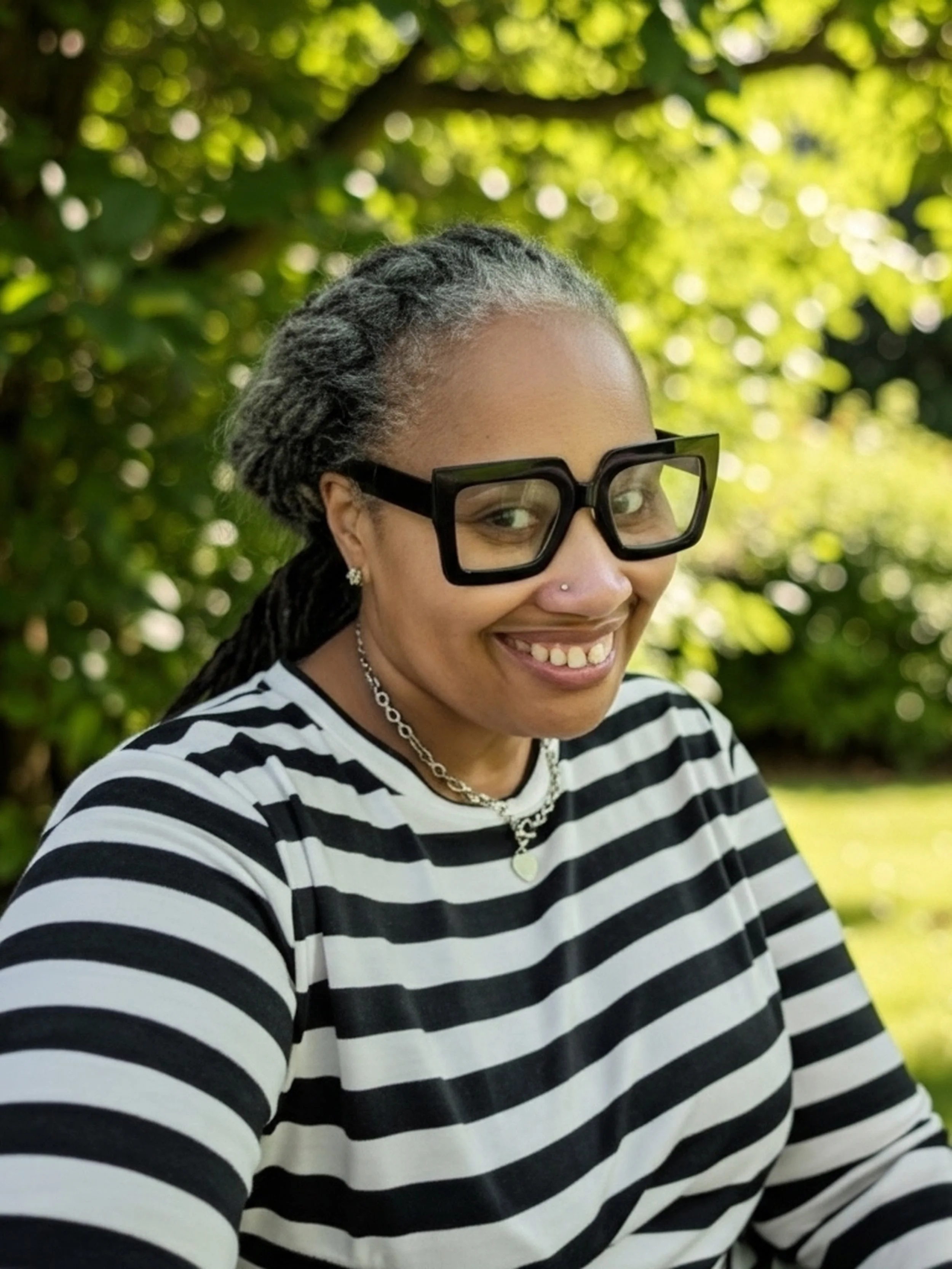 A woman with glasses and dreadlocks smiling outdoors, wearing a black and white striped shirt and jewelry, with green foliage in the background.