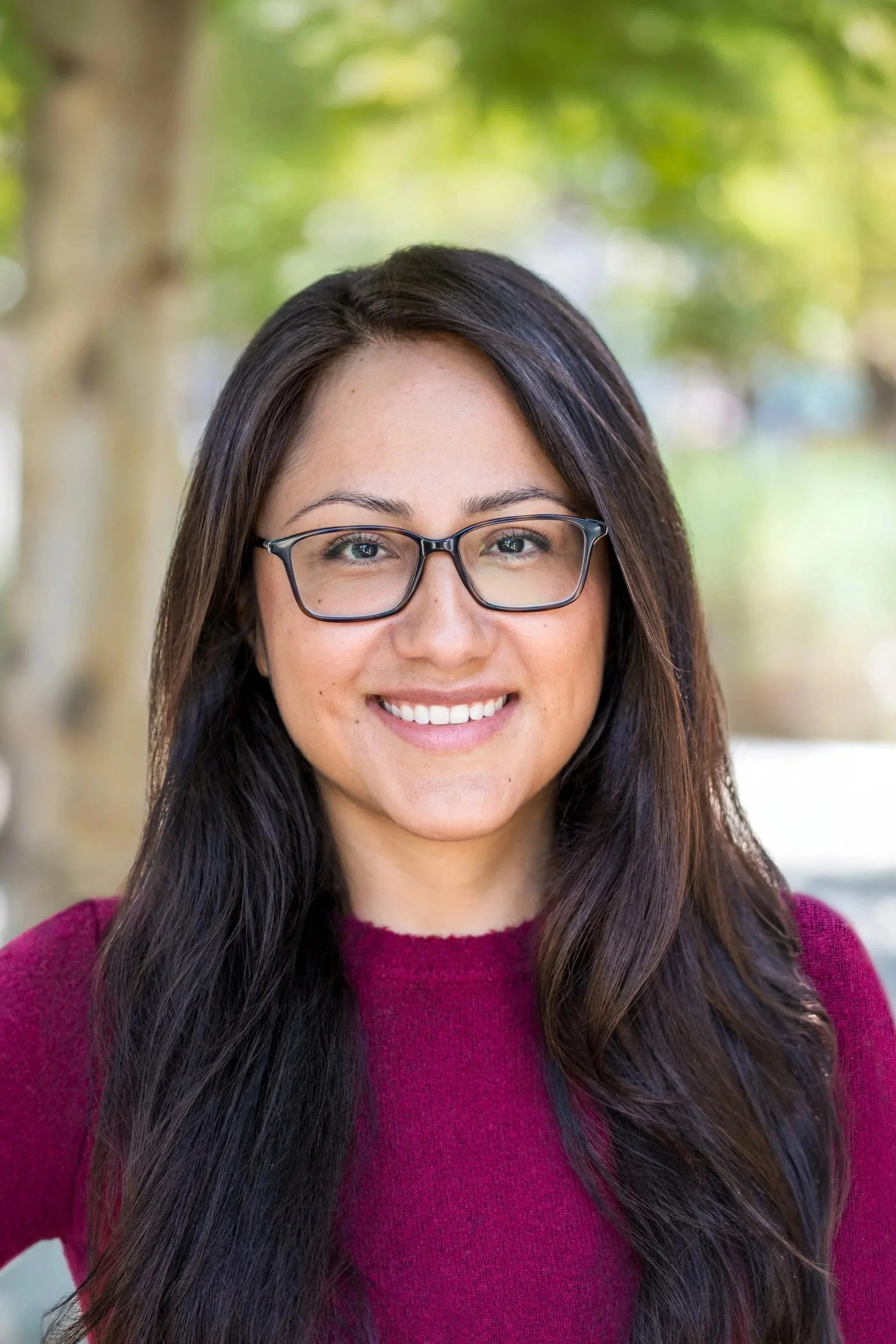 Close-up of a woman with long dark hair, wearing glasses and a maroon sweater, smiling outdoors with blurred green and brown trees in the background.