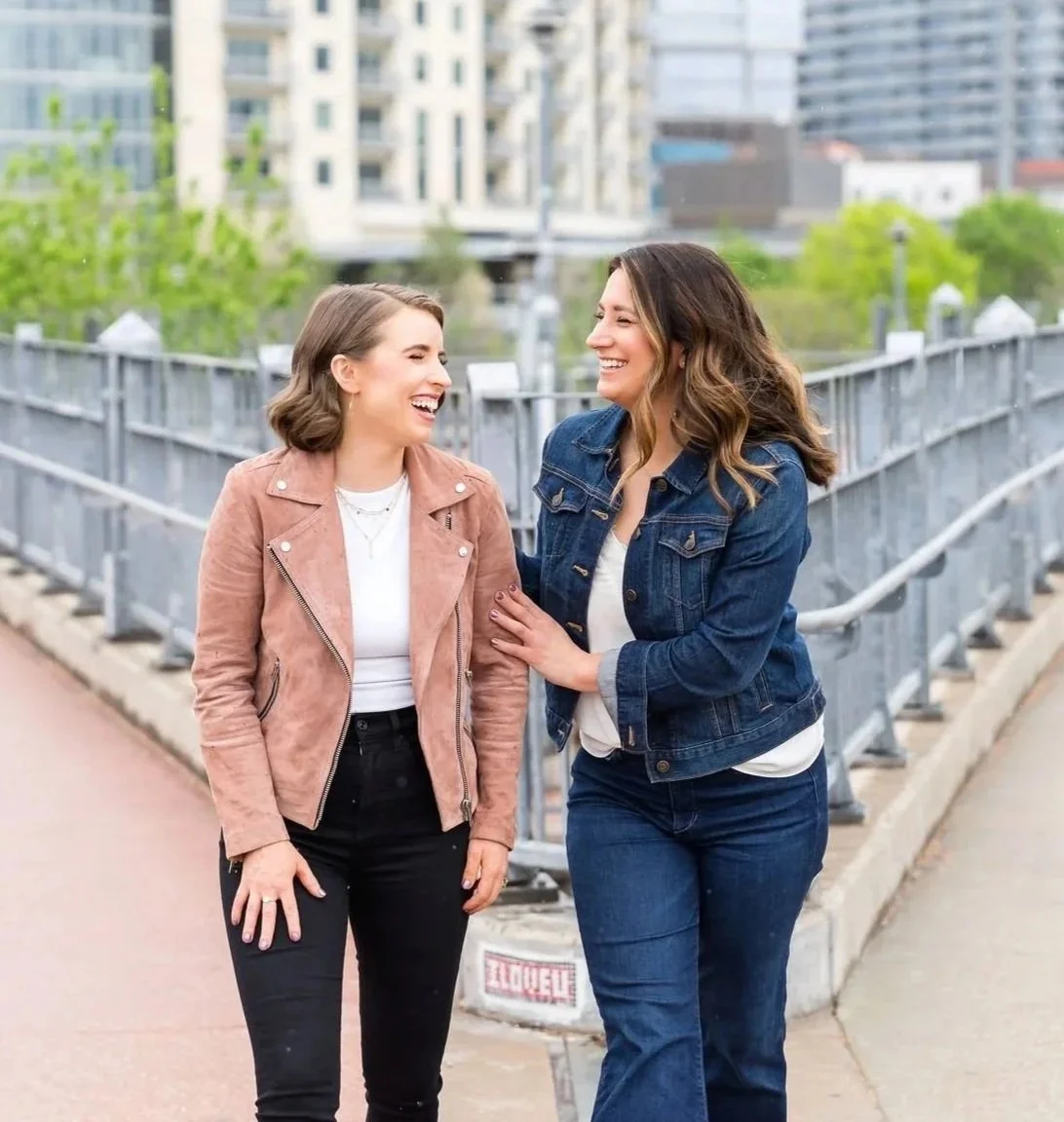 Two women laughing and talking on a city sidewalk, one wearing a pink jacket and the other a denim jacket, with modern buildings and greenery in the background.
