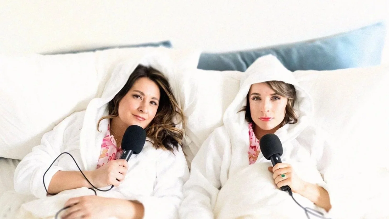 Two women lying in bed, wearing white hooded robes, each holding a black microphone and looking at the camera, with pillows behind them.