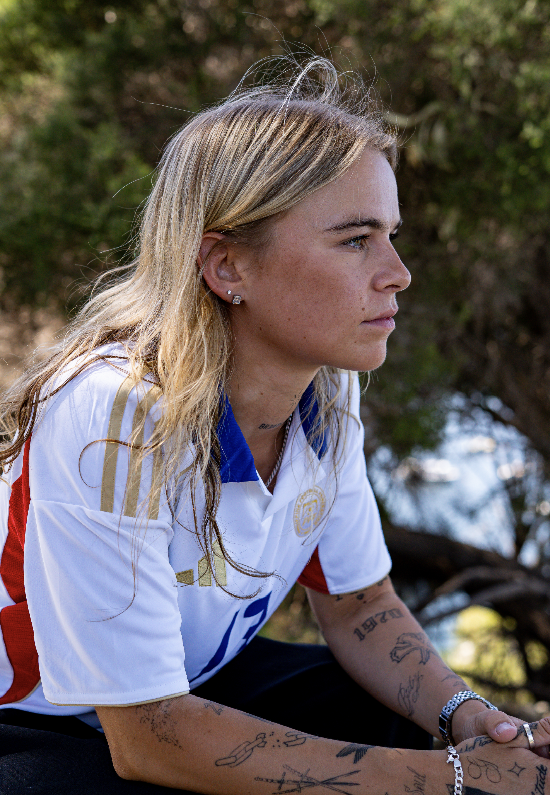 Profile of a woman with long blonde hair and tattoos, wearing a white sports jersey with blue and gold accents, sitting outdoors with trees in the background.
