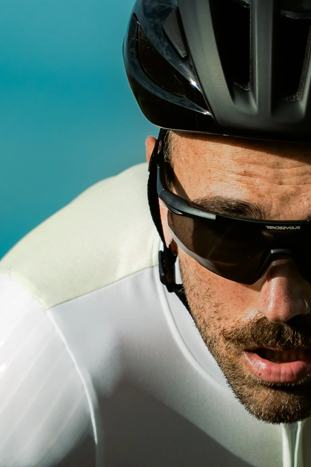 Close-up of a cyclist's face with sunglasses and helmet, captured from a low angle during a ride with a blue sky background.