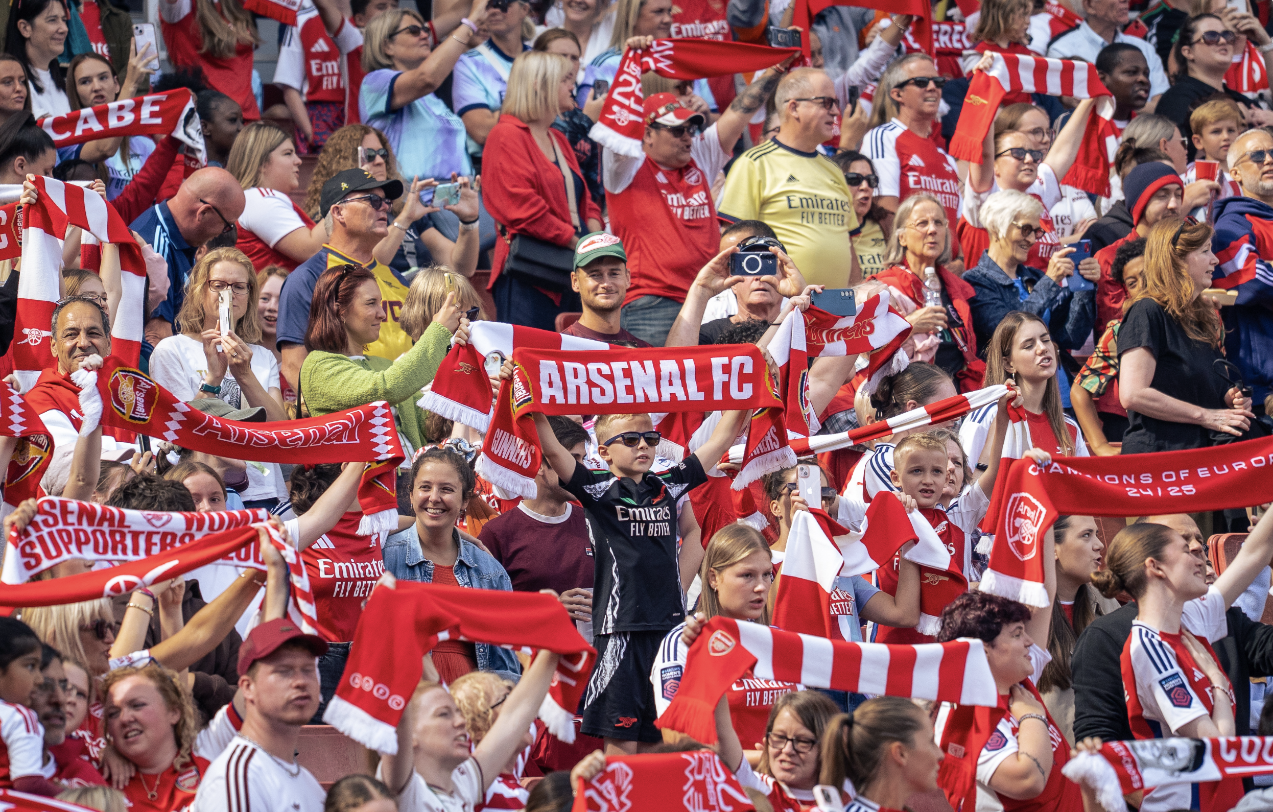 Crowd of soccer fans at a stadium wearing red and white jerseys and scarves supporting Arsenal FC, some taking photos and holding banners.