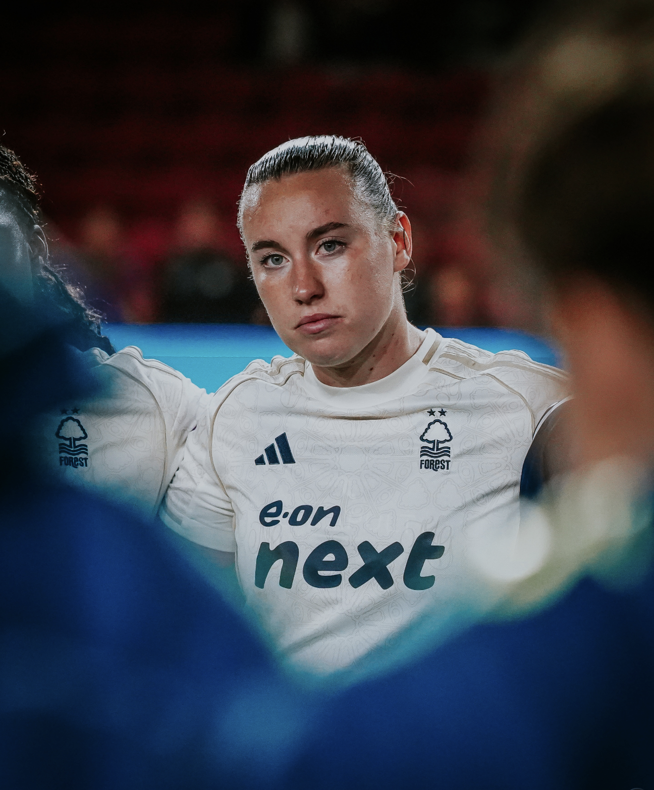 A female athlete in a white sports jersey with blue and black logos, standing with her arms around others in a group, looking serious, with a blurred crowd background.