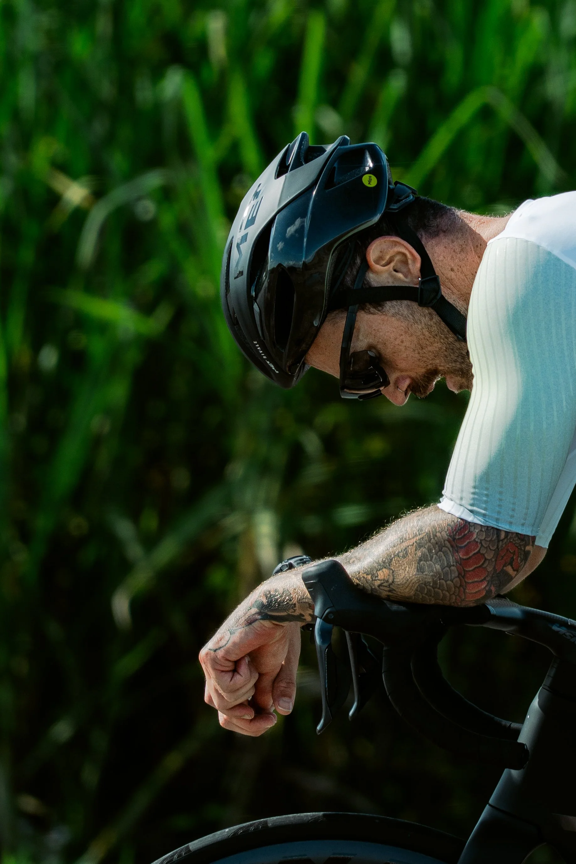 Close-up of a cyclist wearing a helmet and sunglasses, gripping the handlebars of a bike with green foliage in the background.