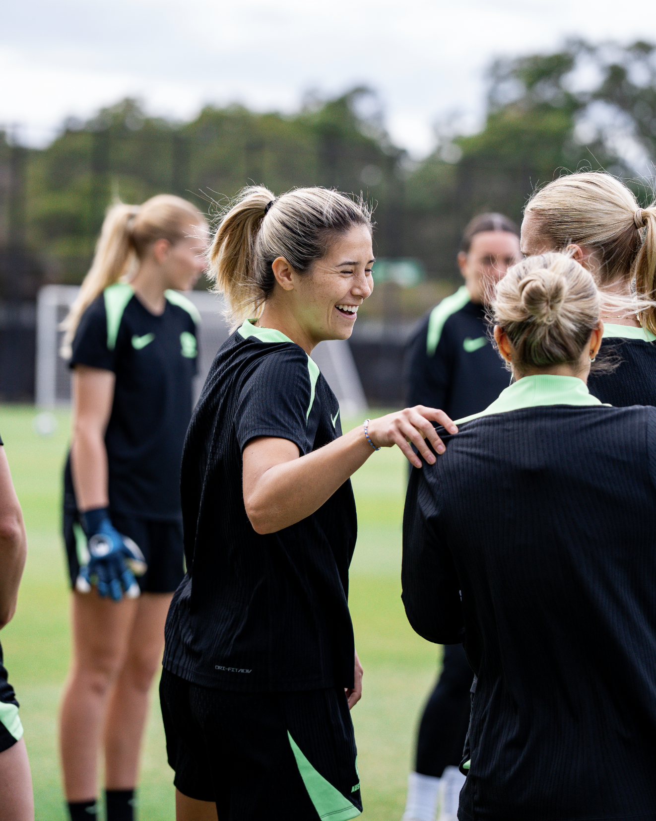A group of female soccer players wearing black and neon green uniforms on a grassy field, smiling and talking with each other.