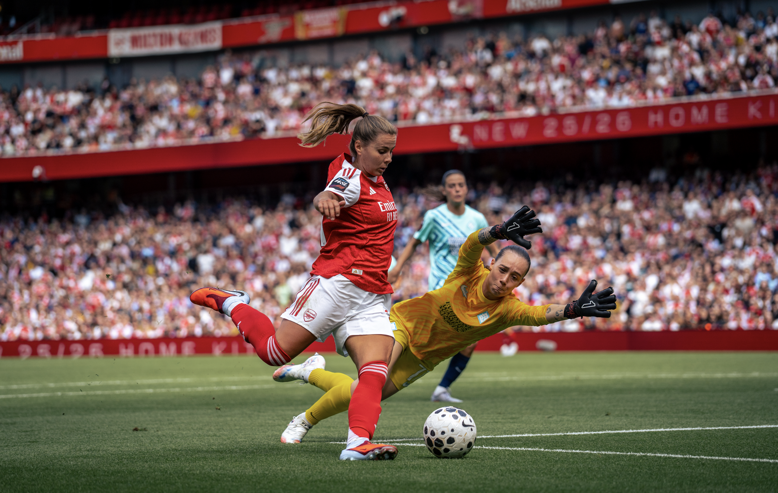 A female soccer player in a red and white uniform kicks a soccer ball while a goalkeeper in a yellow uniform dives to block the shot during a match with a crowded stadium in the background.