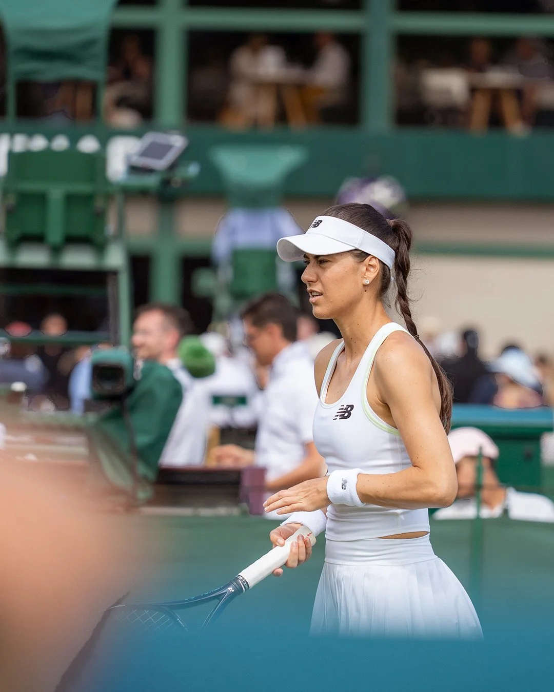 A female tennis player in a white outfit holding a tennis racket on a tennis court, with spectators and officials in the background.