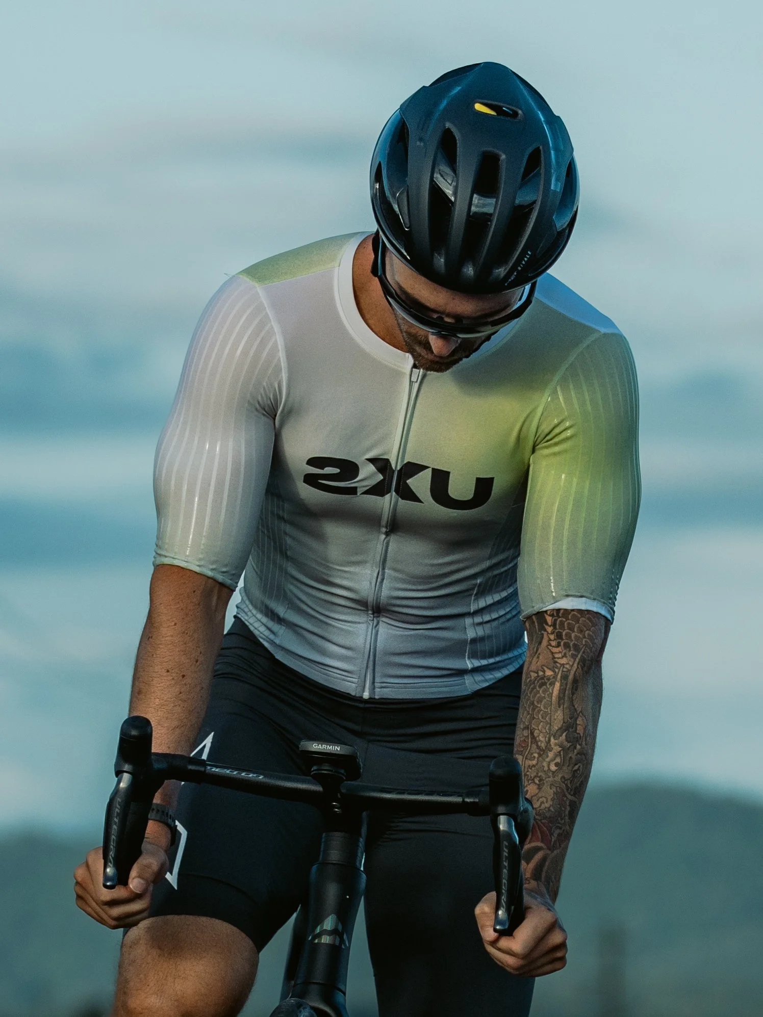 A man wearing a navy cycling helmet, sunglasses, a light-colored cycling jersey with brand logo, and black shorts, riding a bicycle outdoors with a blurred sky background.