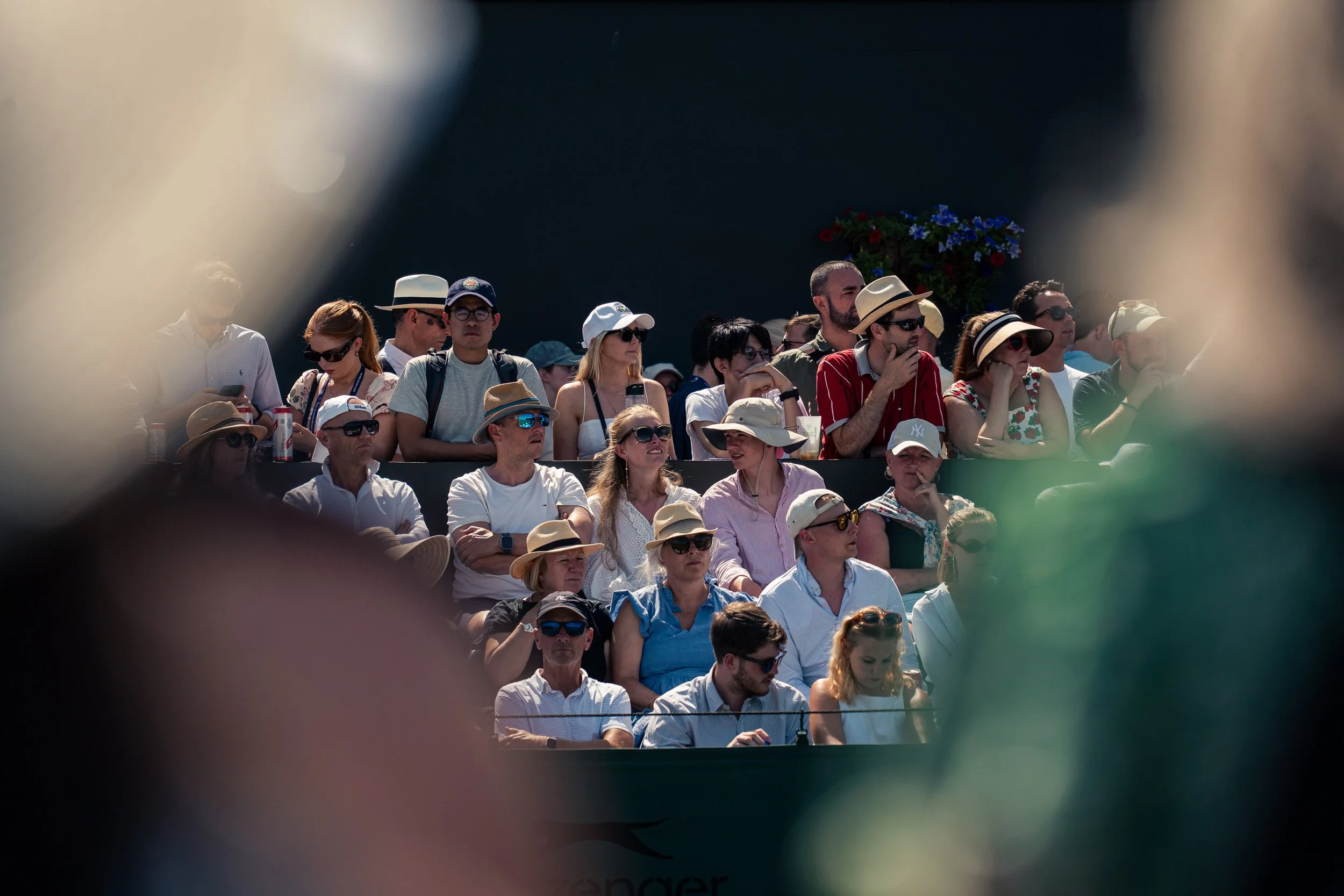 A crowd of people seated outdoors, some wearing hats and sunglasses, seen through a small opening or frame.
