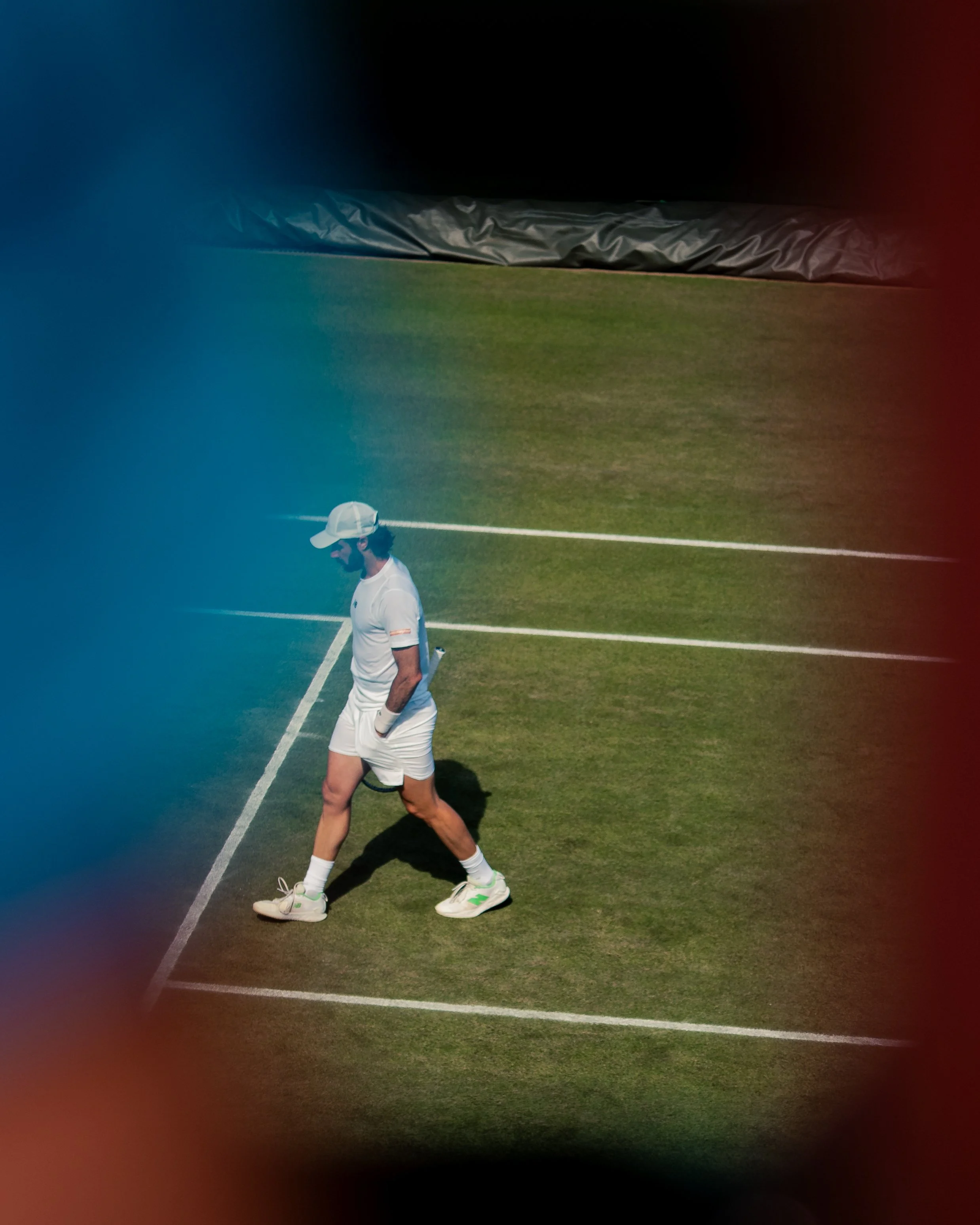 A person in a white sports outfit and cap walking on a tennis court at night, with part of the court, a net, and a dark background visible.