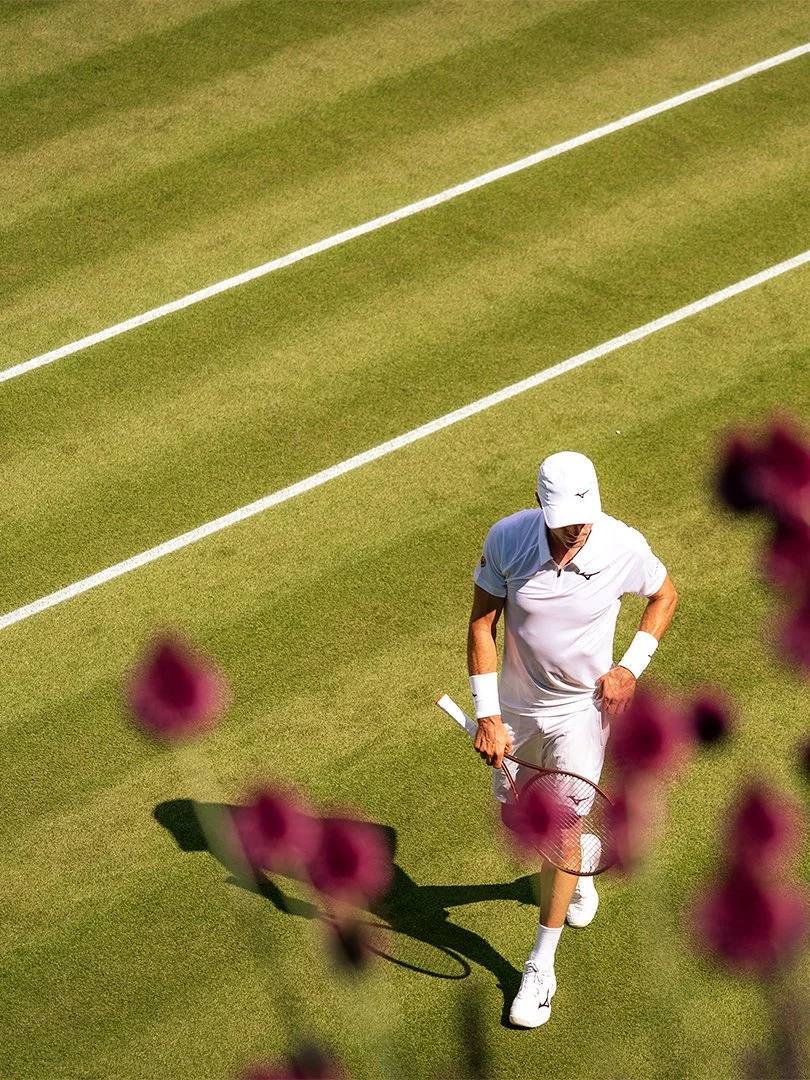 A golfer dressed in white holding a tennis racket walking on a green grass tennis court with white boundary lines, partially obscured by pink flowers in the foreground.