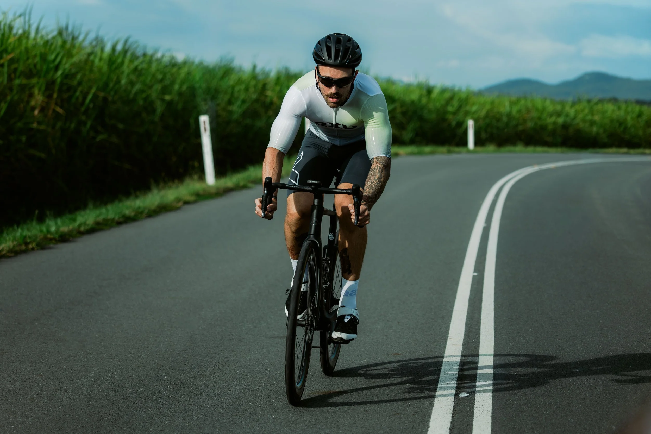A man riding a black road bicycle on a curved rural road, wearing a black helmet, sunglasses, and a cycling outfit.