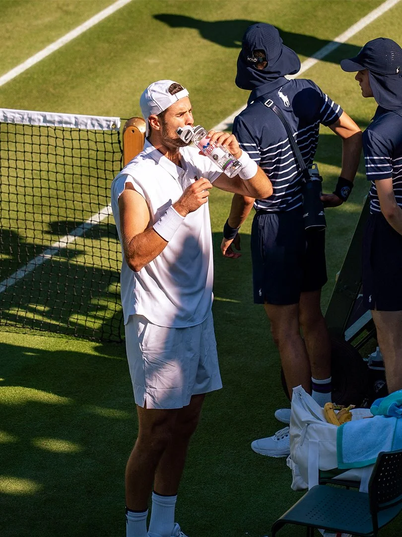 A male tennis player in a white outfit drinking water from a water bottle on a grass court, with a tennis net and two other players in dark blue outfits nearby.