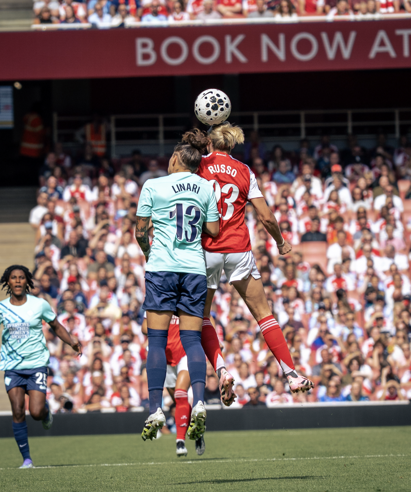 Two female soccer players, one in a light blue jersey numbered 13 and the other in a red jersey numbered 23, jump to head a soccer ball during a match. The crowd watches from the stands.