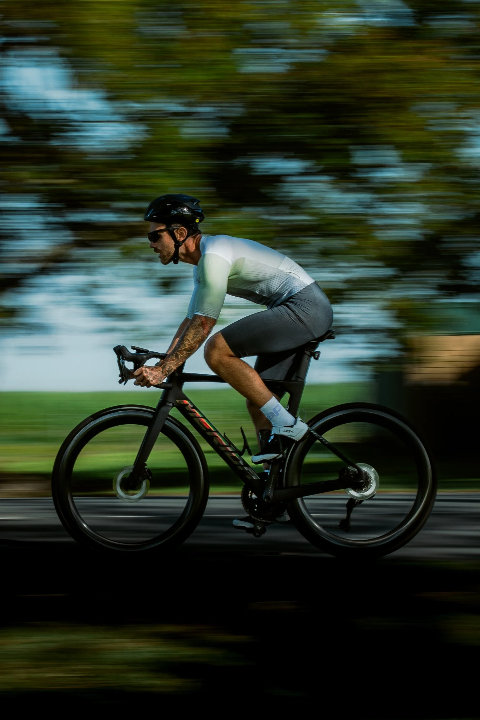 A man riding a black bicycle wearing a helmet, sunglasses, a white and gray athletic shirt, black shorts, and cycling shoes across a blurred background of trees and sky.
