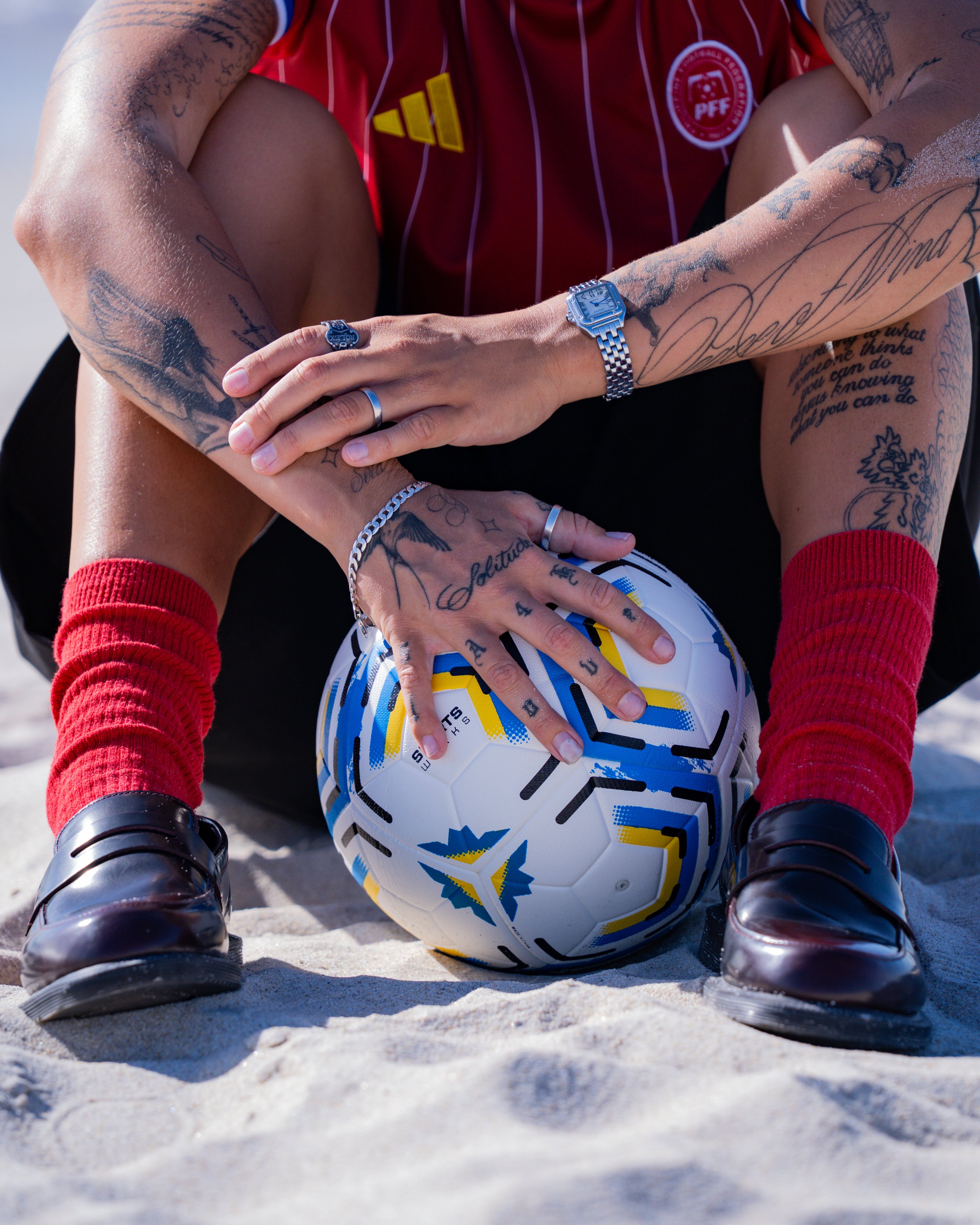 Close-up of a soccer ball on sandy ground between two children’s legs, with one hand resting on the ball. The children are wearing red gloves, black shoes, and bright clothing. The children have tattoos on their arms.