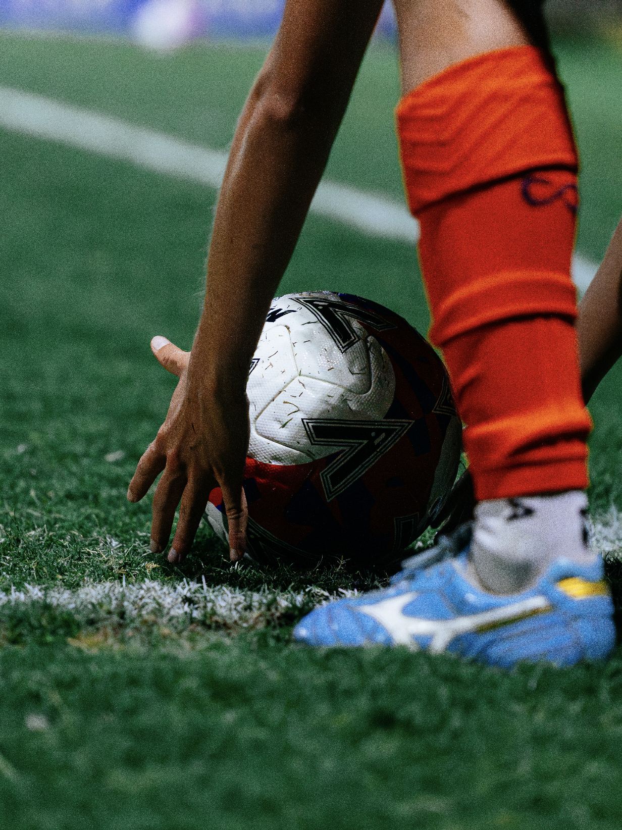 A soccer player is preparing to kick a white soccer ball on a green field, wearing orange shorts and white soccer cleats.