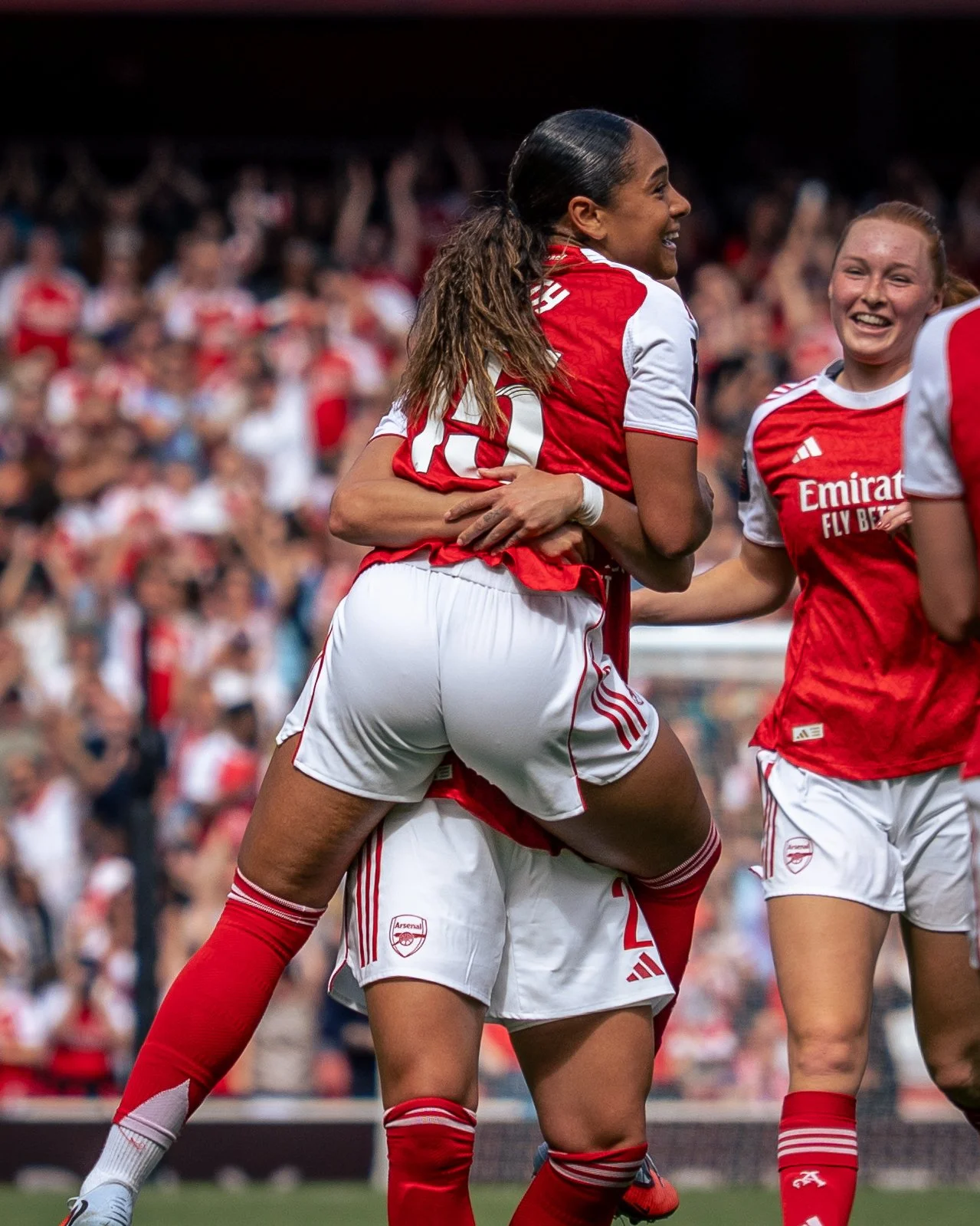 Arsenal female soccer players celebrating on the field, wearing red and white uniforms, in a stadium with a crowd in the background.