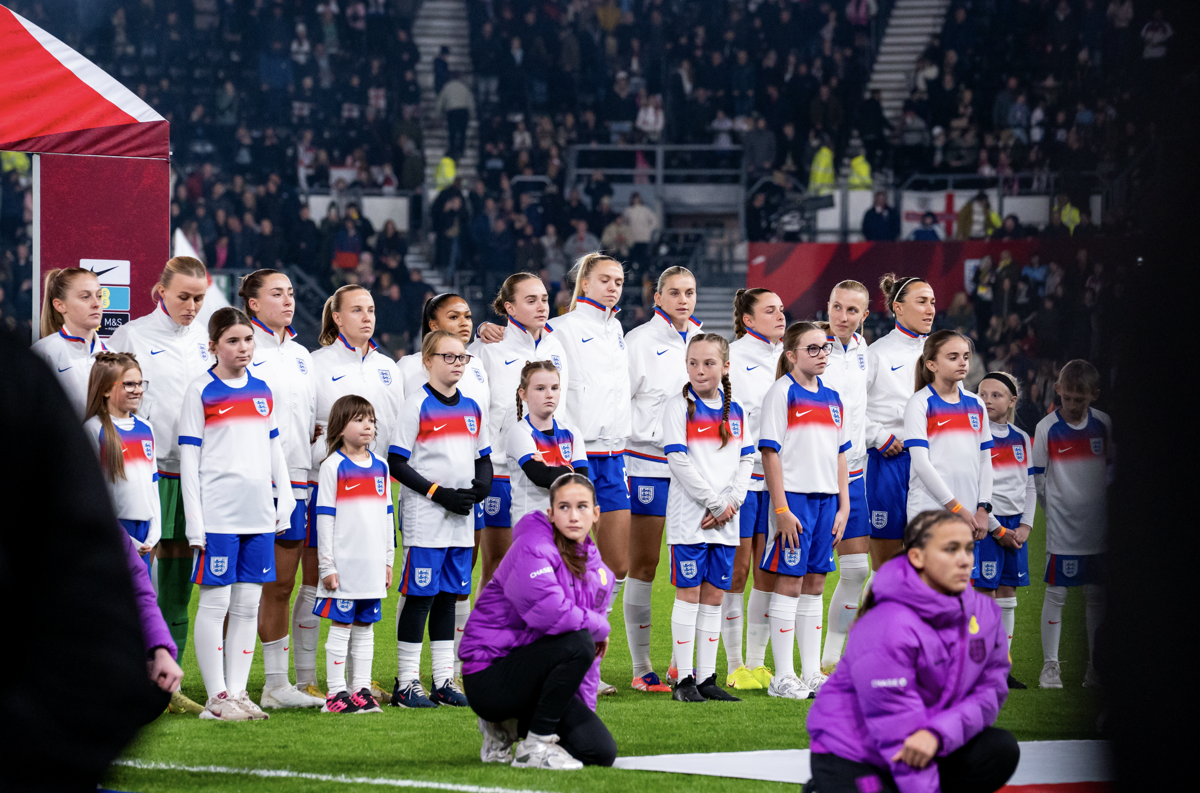 A group of female soccer players and young girls standing on the field, some kneeling and others standing, during a pre-match ceremony at a stadium with spectators in the background.