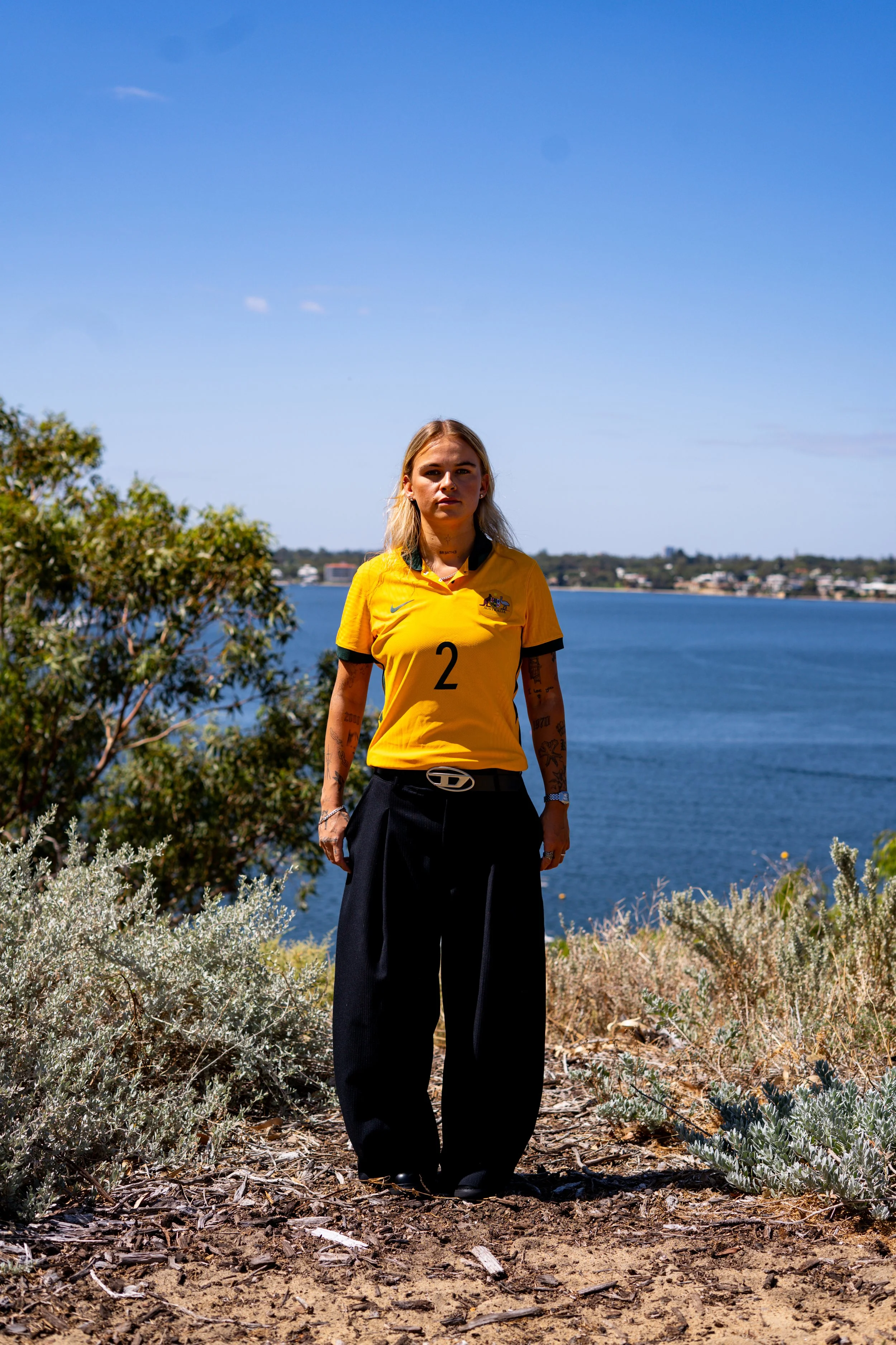 Young woman with tattoos wearing a yellow sports jersey and black pants standing outdoors near water and trees on a sunny day.