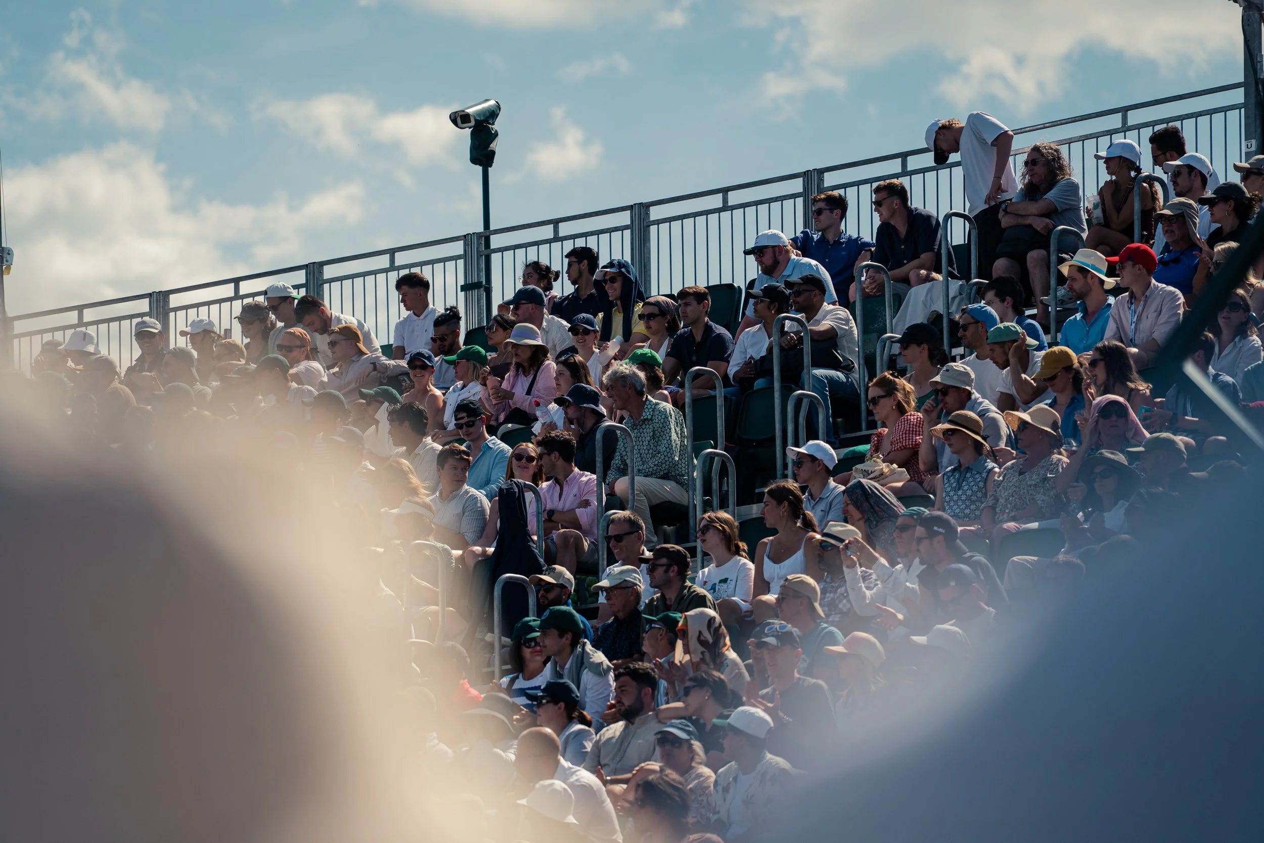 Crowd of people seated outdoors in tiered seating, some wearing hats and sunglasses, with a blue sky and clouds in the background.
