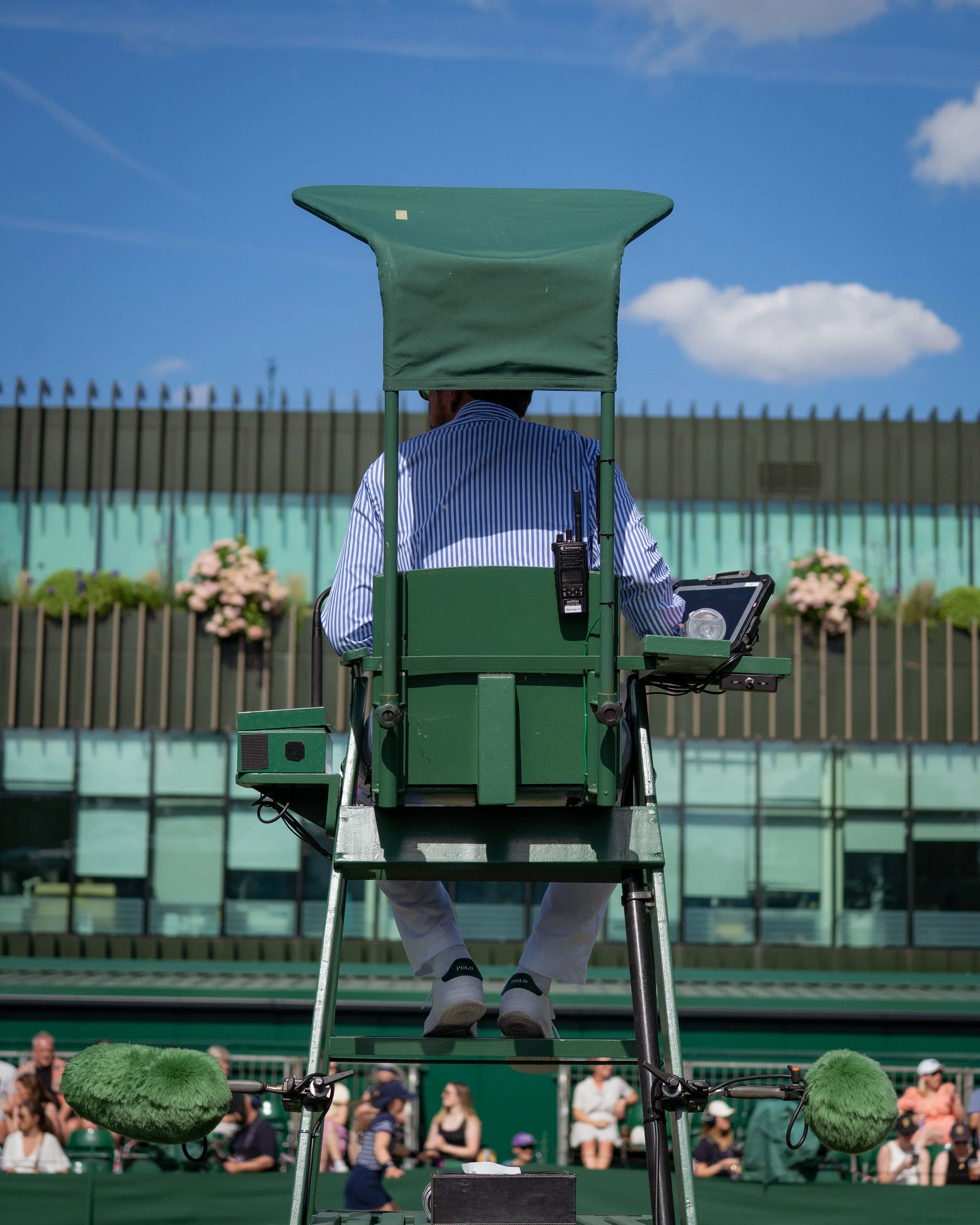 Umpire sitting on a chair behind a tennis court, overlooking the game, with crowd watching in the background.