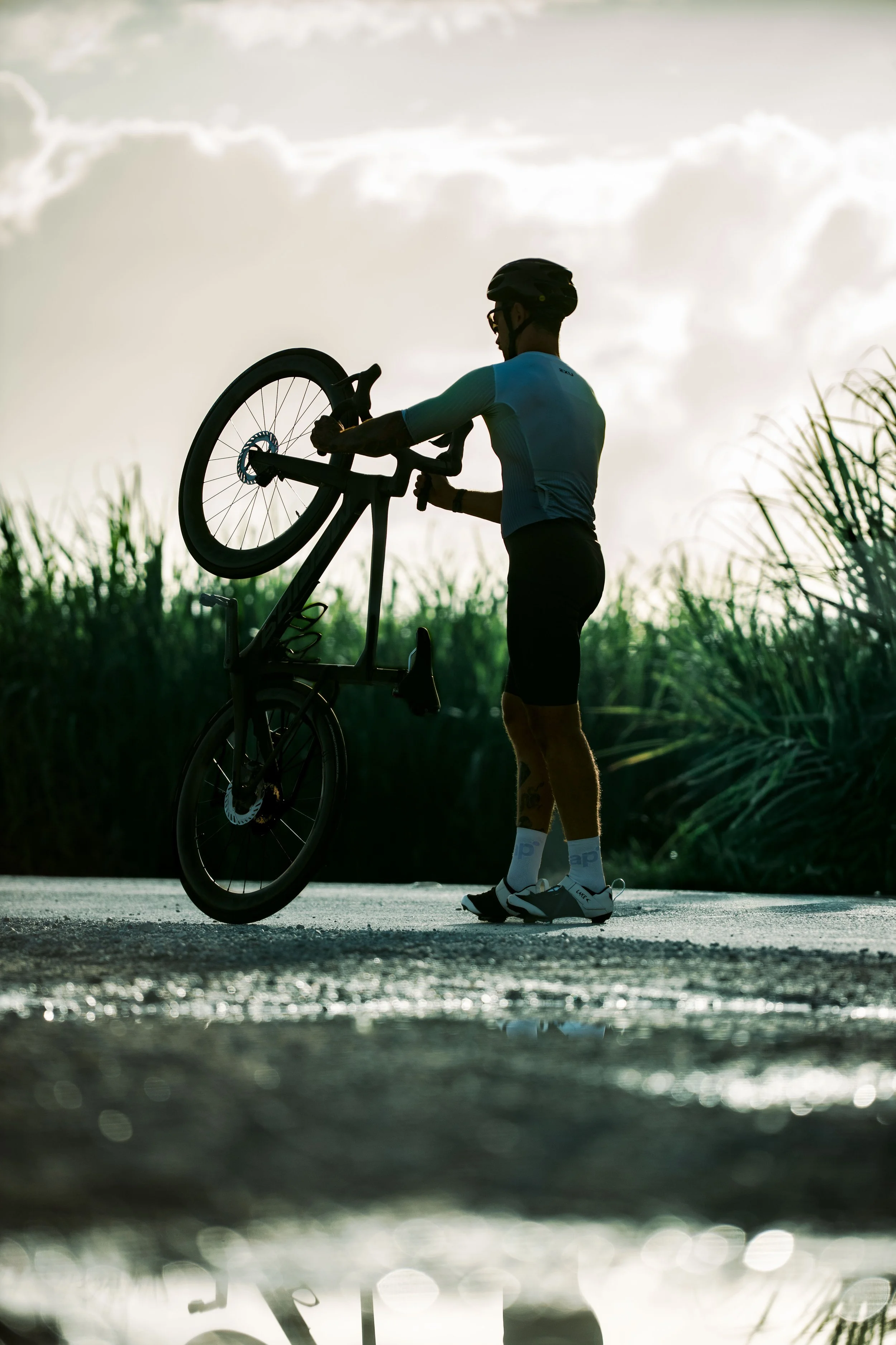 A cyclist in a blue jersey and a black helmet hands off his bike on a path near a body of water during sunset, with tall grasses and a cloudy sky in the background.