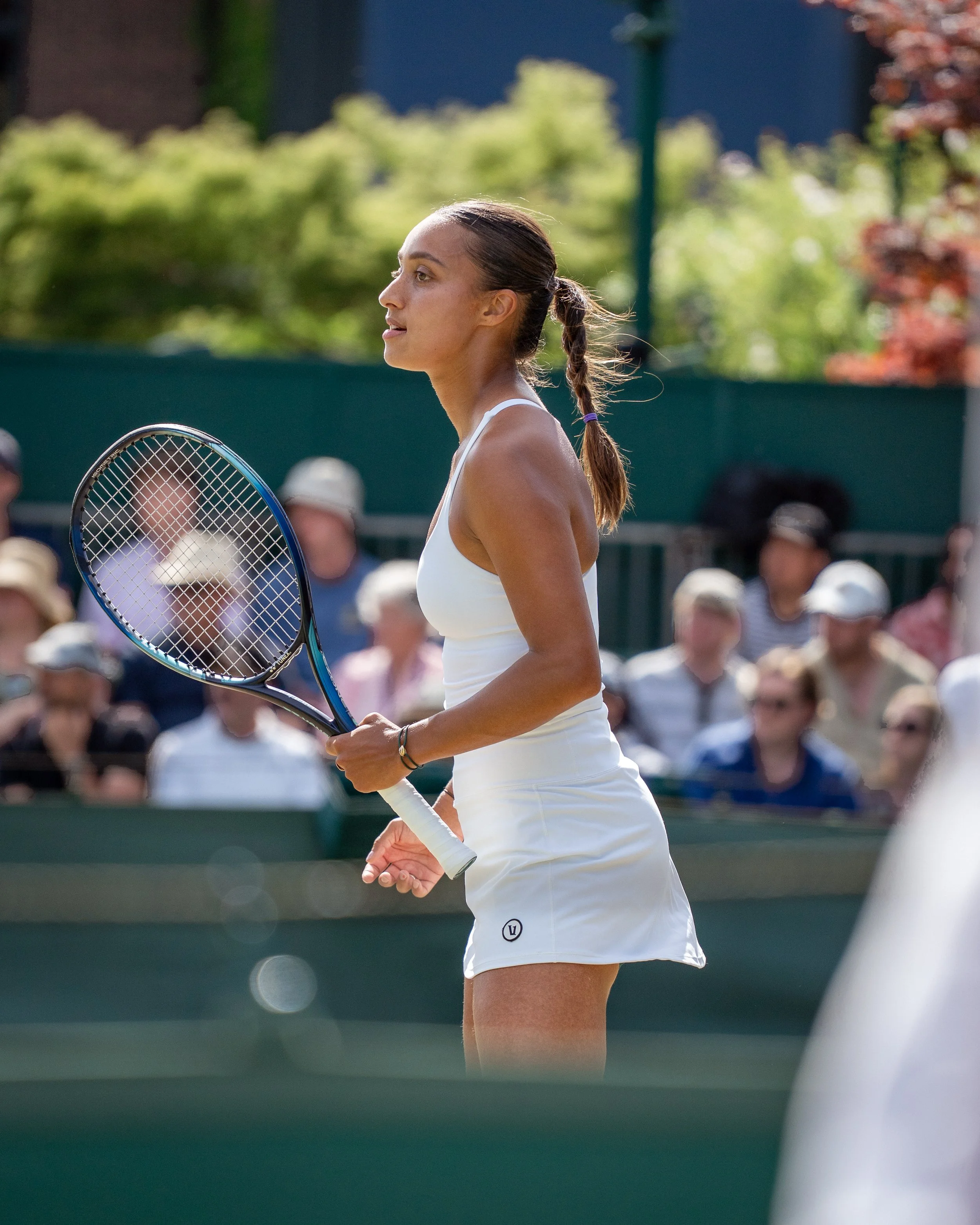 Side view of a female tennis player wearing a white tank top and skirt, holding a tennis racket, during a match on an outdoor court with spectators in the background.