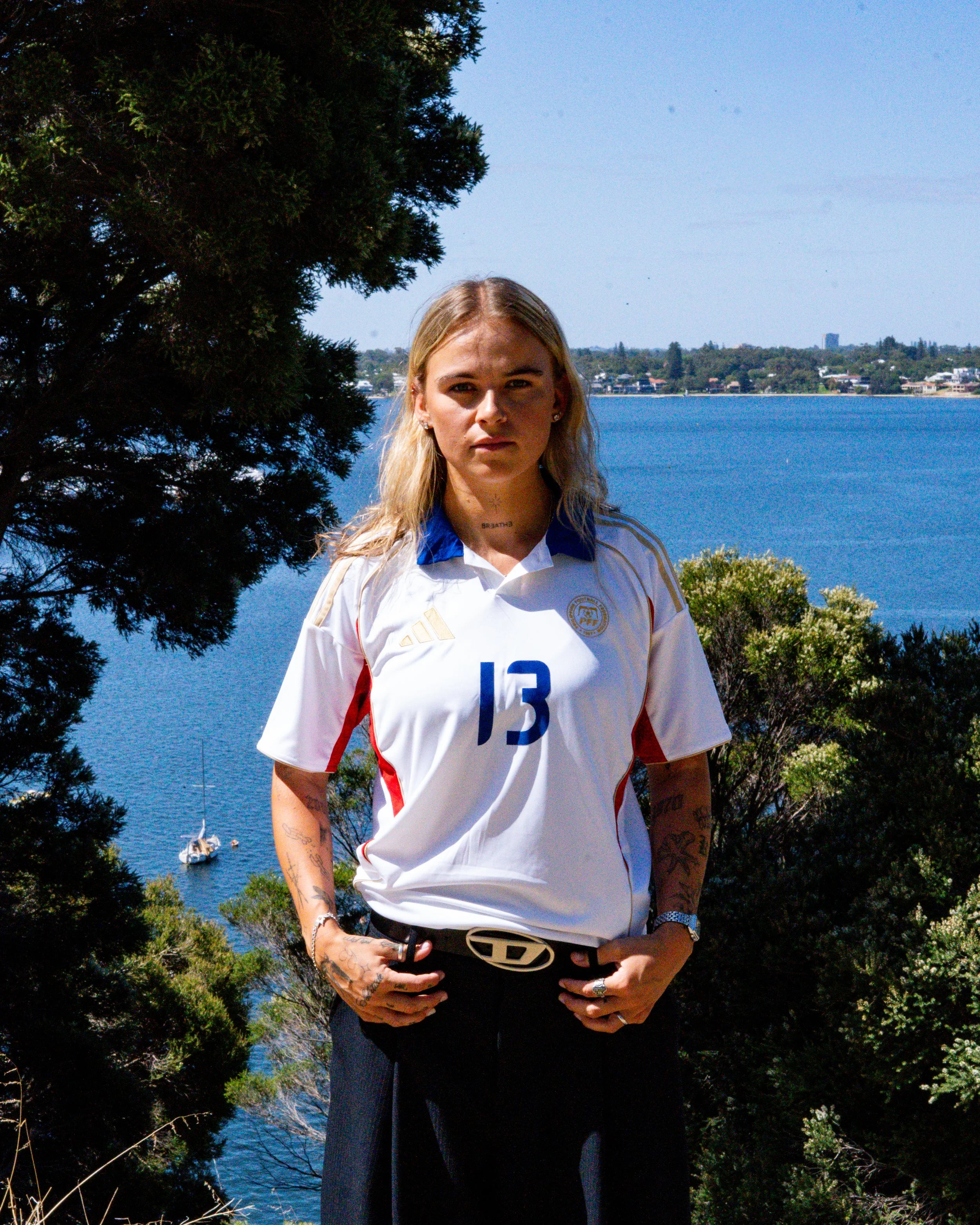 A woman with blonde hair and tattoos on her arms posing outdoors near water with trees and boats in the background.