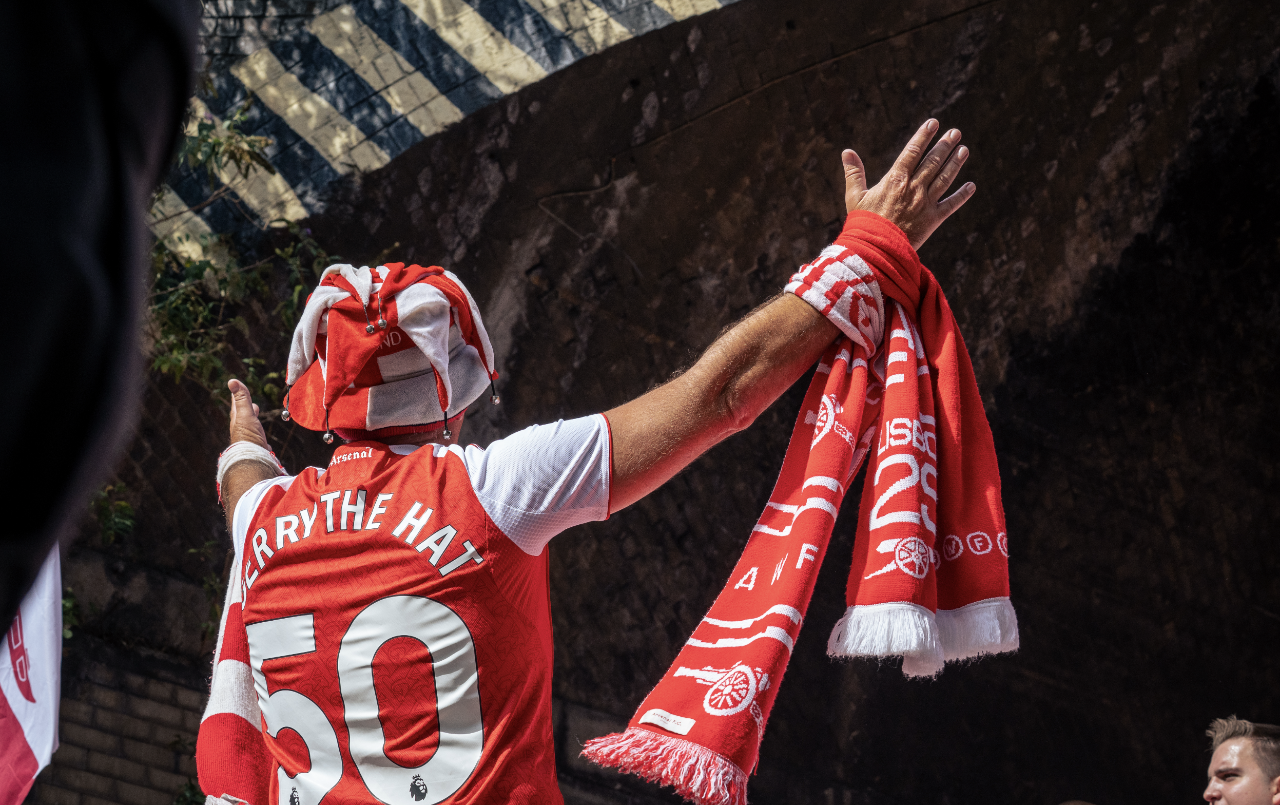 A person wearing an Arsenal football jersey and a matching hat with jingle bells, holding a red and white scarf with team logos, raising their hand in celebration against a brick wall background.