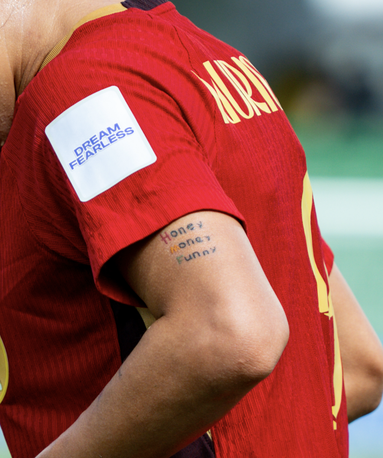 A close-up of a person wearing a red sports jersey with yellow accents. There is a patch on the sleeve that reads "DREAM FEARLESS" in blue text. The person's arm has a tattoo that says "Honey Honey Funny" in colorful ink.