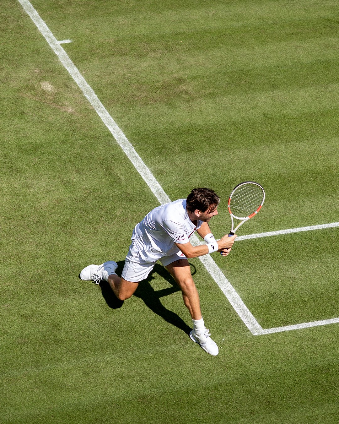 A tennis player in white clothes is on his knees on a grass court, holding a tennis racket and preparing to hit a ball.