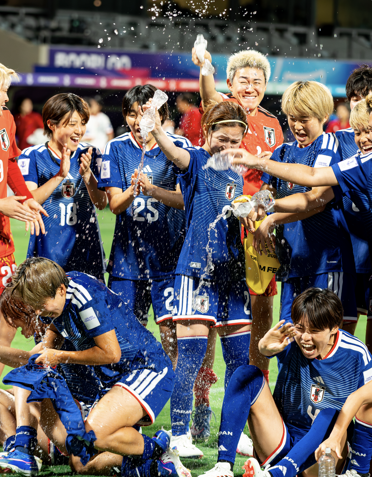 Japanese women's soccer team celebrating on the field, pouring water over each other, wearing blue jerseys with the Japan Football Association emblem, after winning a match.