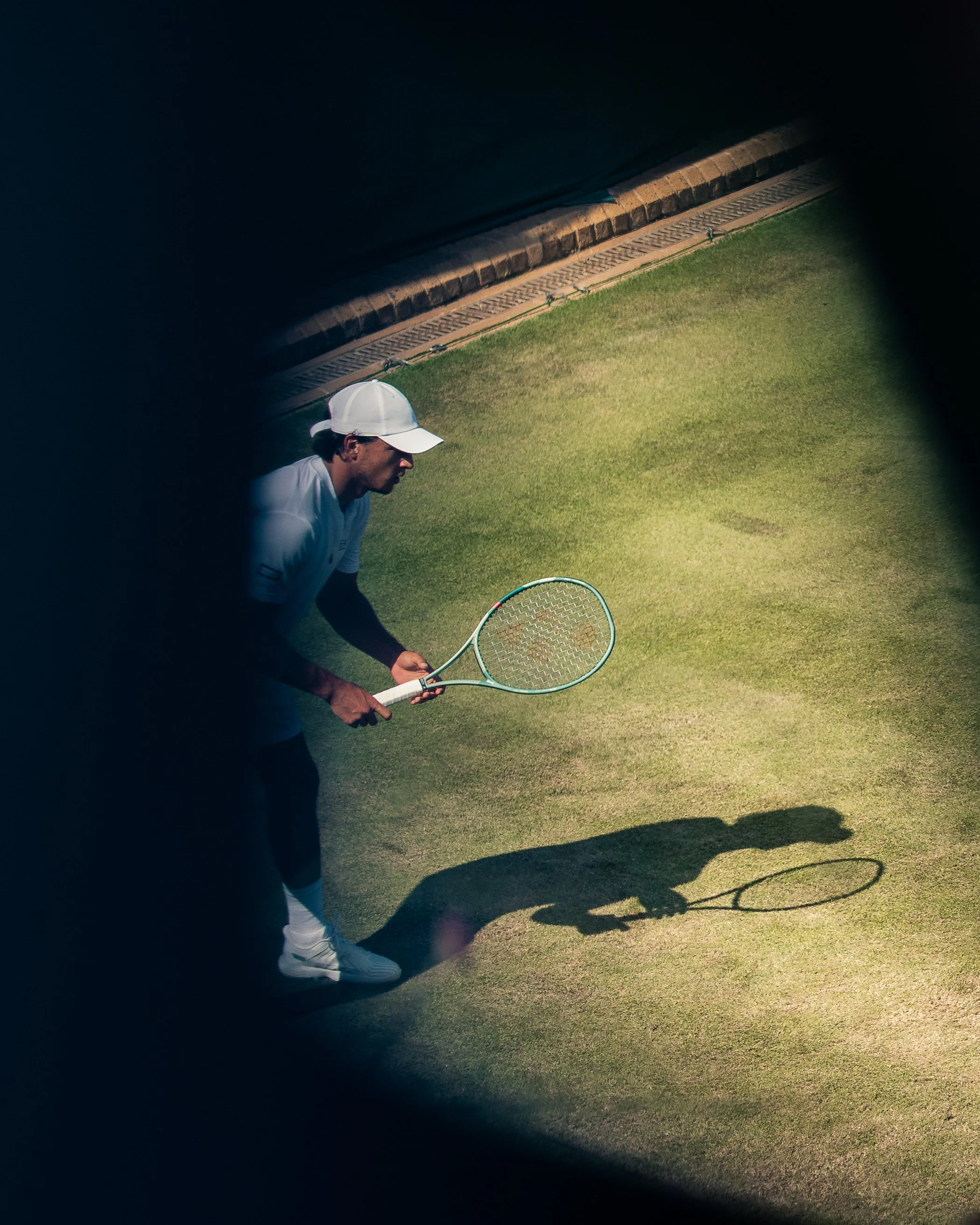 A man dressed in tennis attire, wearing a white cap, bowing on a tennis court, with sunlight casting shadows.