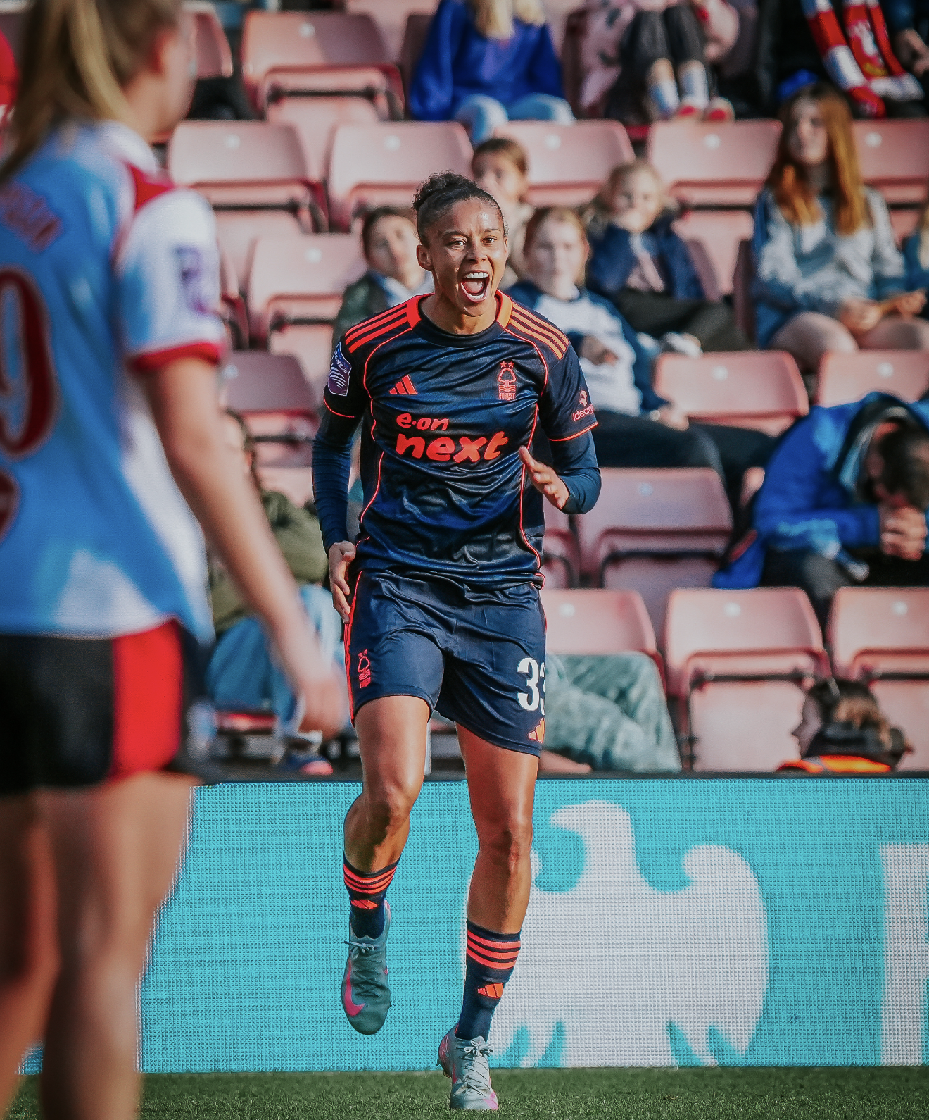 A female soccer player in a dark blue uniform with red accents, running and celebrating on the field during a match, with spectators sitting in the stadium seats behind her.