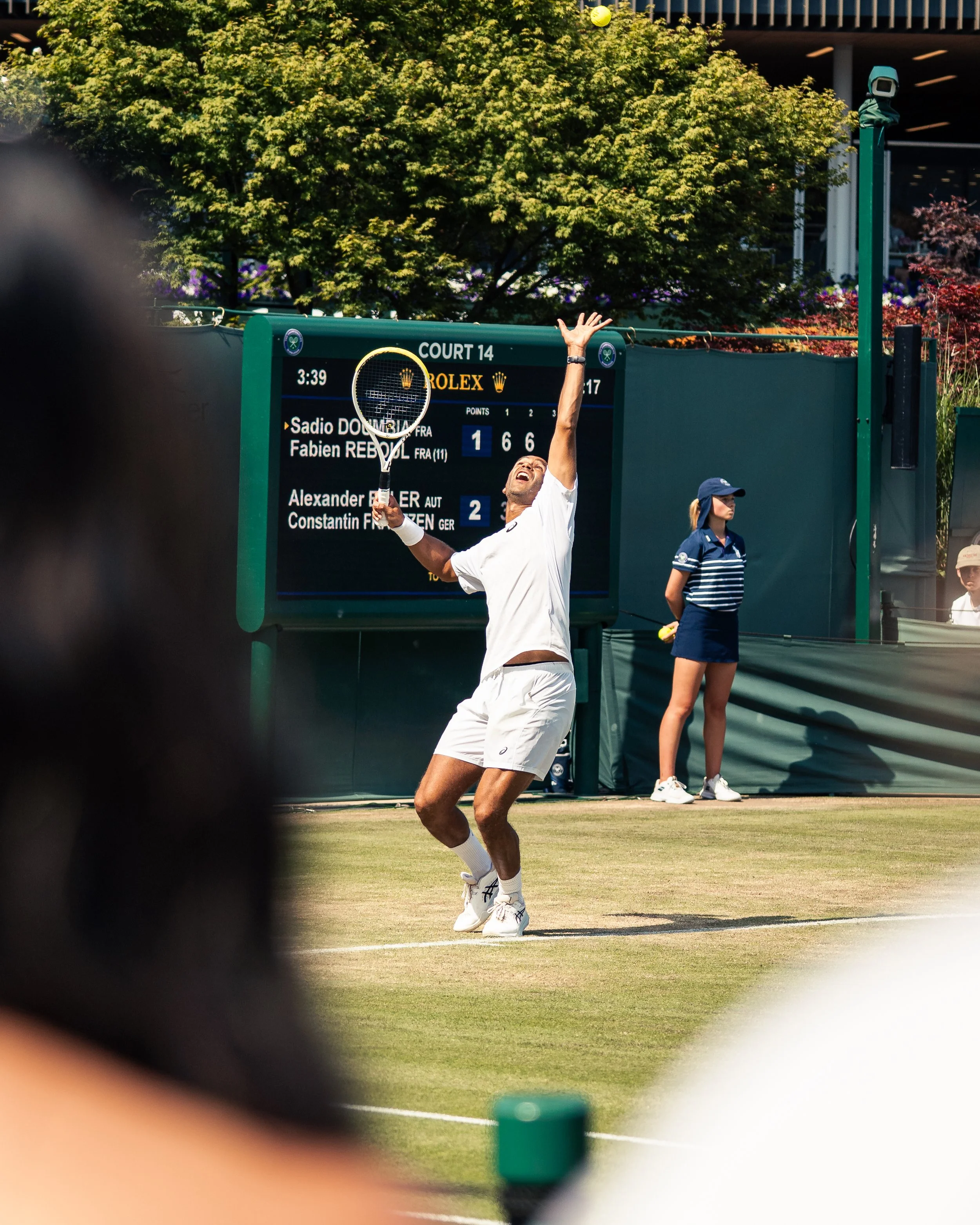 A tennis player in a white shirt and shorts is preparing to hit a serve on a grass court. The scoreboard behind him shows a score of 1-6, 6-6 in the match. An official and a ball girl are also visible near the court.