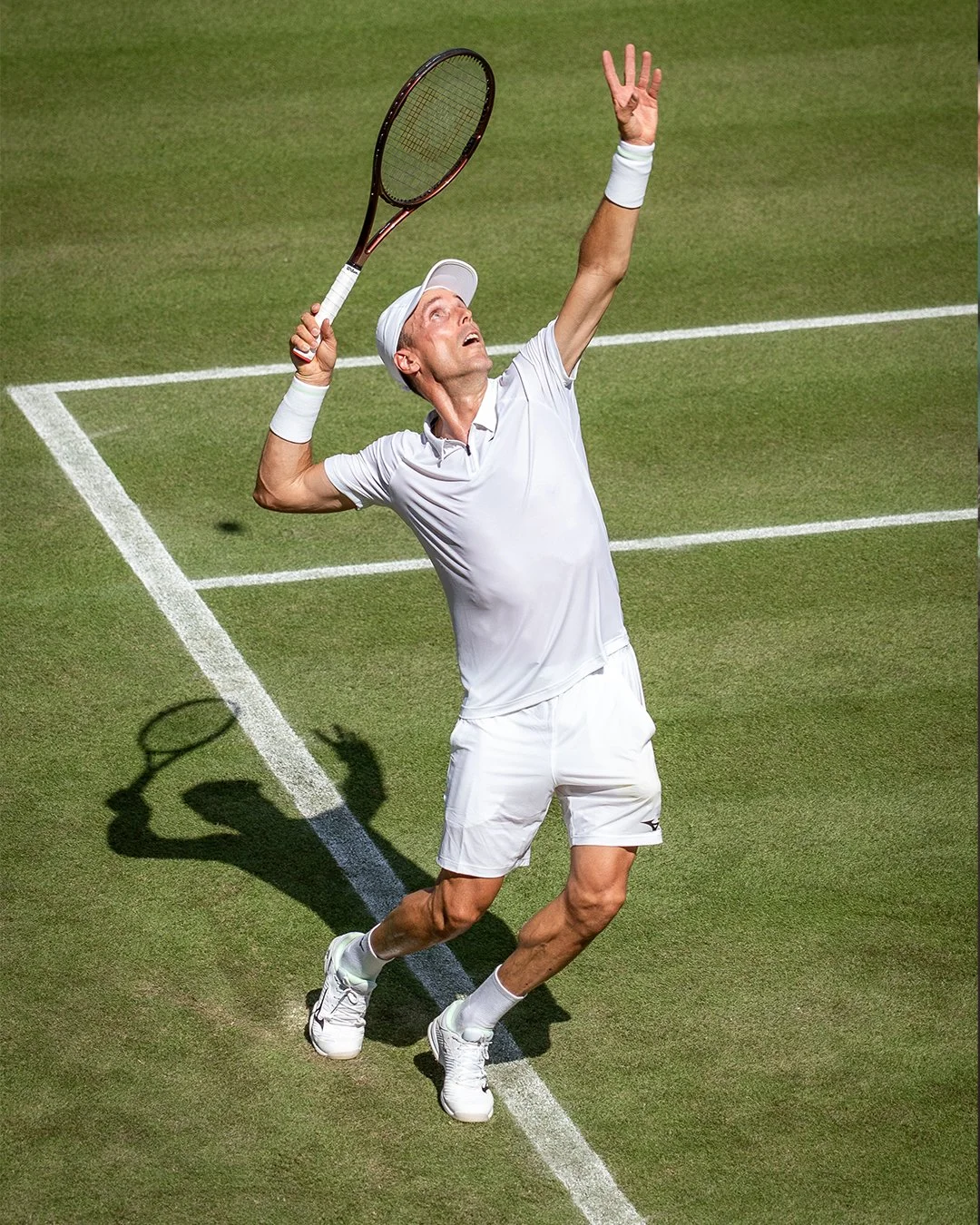 A male tennis player in white athletic wear, including a shirt, shorts, and tennis shoes, is on a grass court preparing to hit a tennis ball with a racket, casting a shadow on the ground.