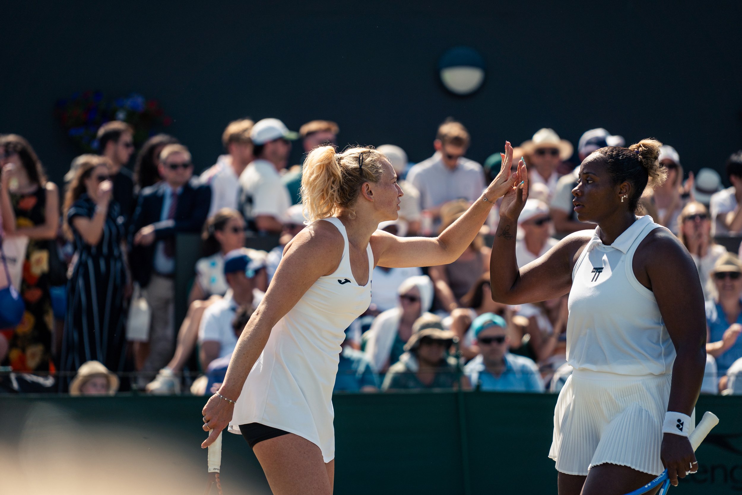 Two female tennis players, one with blonde hair and the other with dark hair, greeting each other on a tennis court amid a crowd of spectators.
