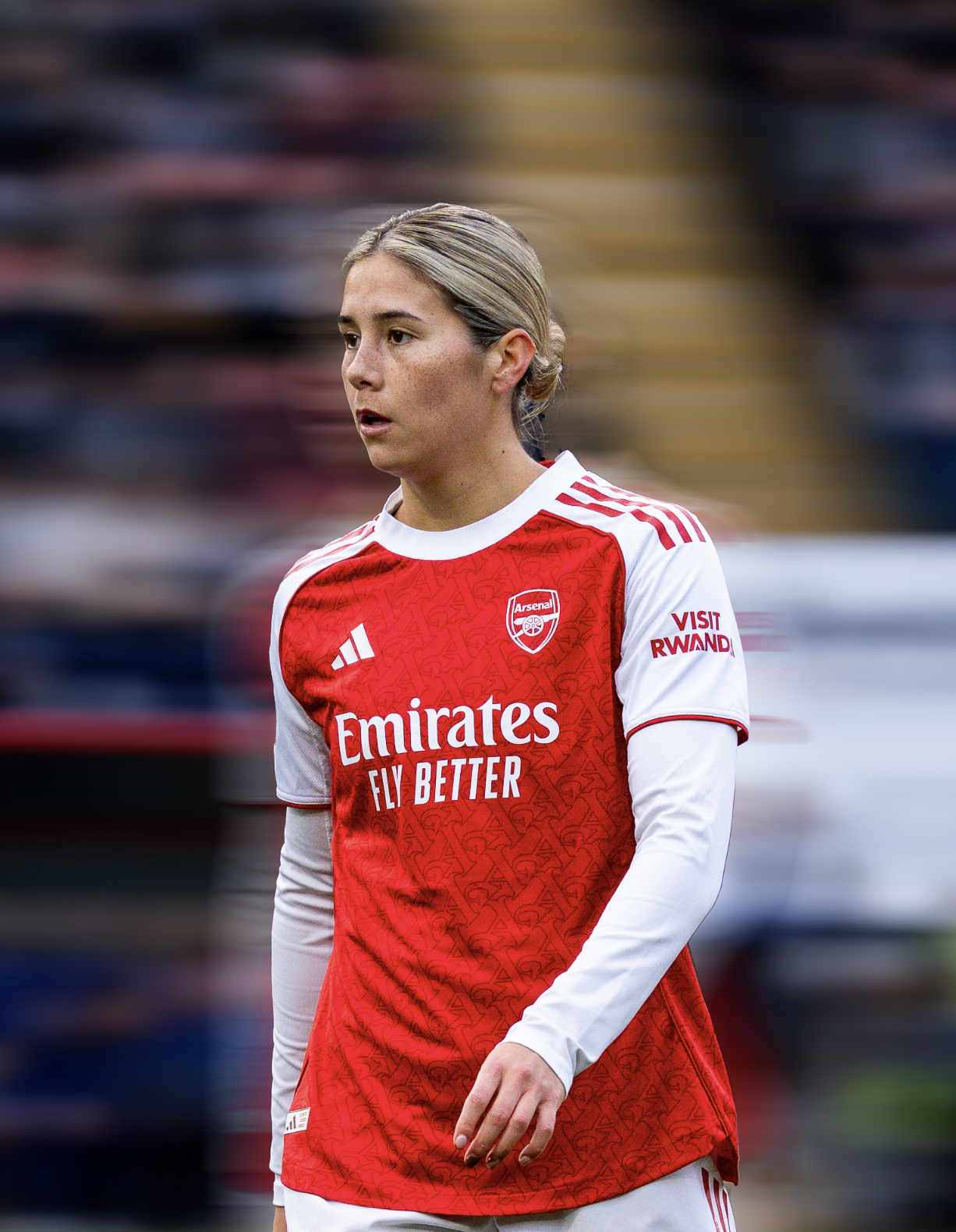 A female soccer player wearing an Arsenal jersey with the Arsenal logo, and sponsor logos, standing on the field with a blurred crowd in the background.