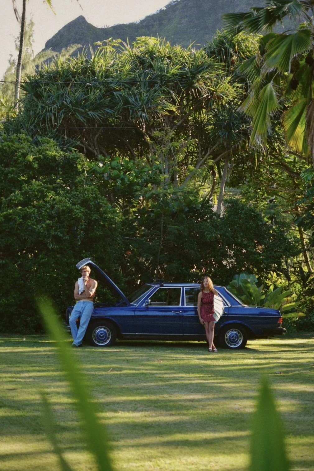A man and woman standing next to a dark blue vintage car on a grassy field, surrounded by lush green tropical trees with a mountain in the background.