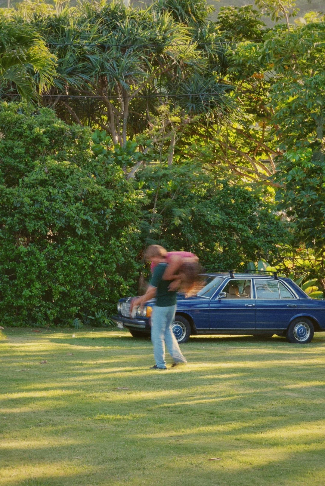A woman in casual clothes stands on a well-manicured lawn, bending forward with her hair flying, in front of a blue vintage Mercedes-Benz car, surrounded by lush green bushes and tall trees in sunlight.