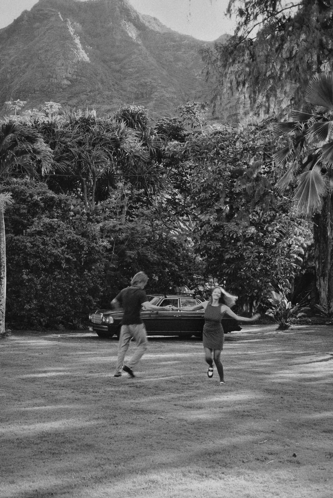 A black and white image of a couple dancing outdoors on a grassy area, with a vintage car parked behind them and lush trees and mountains in the background.