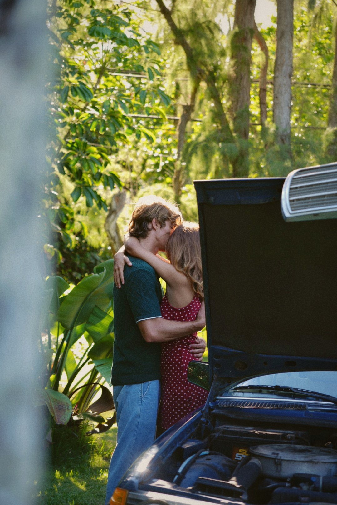 A couple embracing tenderly outdoors in a lush, green setting near a car with its hood open.