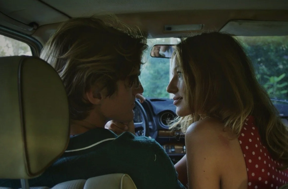 A young couple sitting close together in the front seat of a vehicle, looking at each other with the man lying back and the woman leaning in.