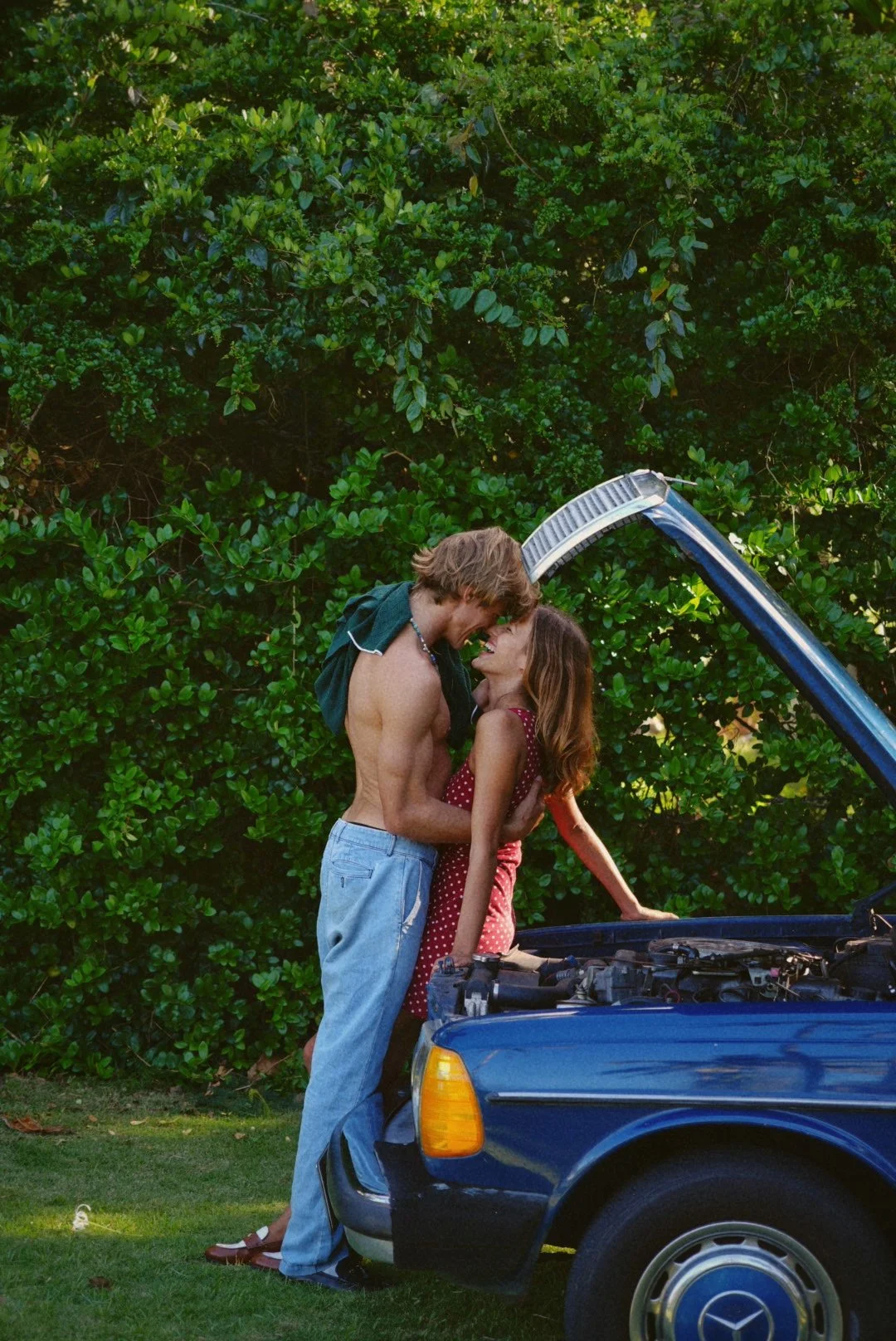 A man and woman are standing close together next to a blue car with its hood open, sharing an affectionate moment outdoors with green shrubbery in the background.