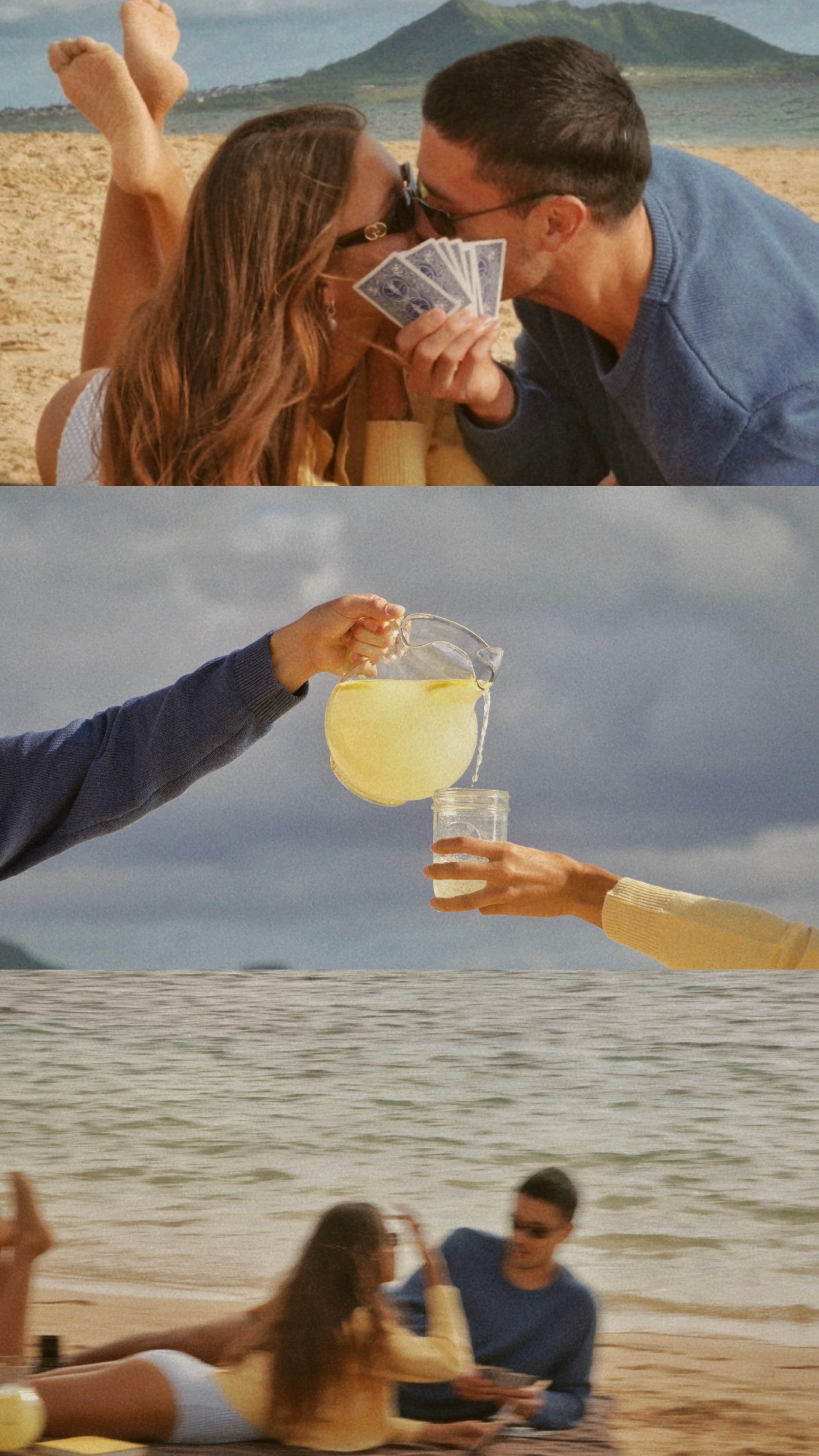 People at the beach enjoying a romantic moment, with some holding cards and a pitcher of lemonade, and others lounging on the sand near the ocean with mountains in the background.