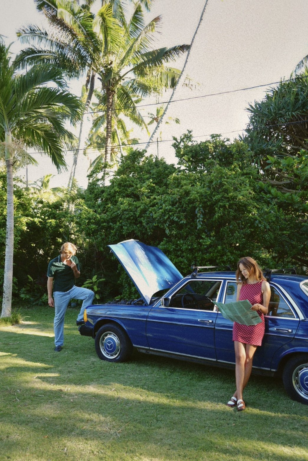 A young man and woman are inspecting a vintage blue car outdoors in a lush, tropical setting with tall palm trees. The woman is standing next to the car, reading a map, while the man is looking under the open hood.